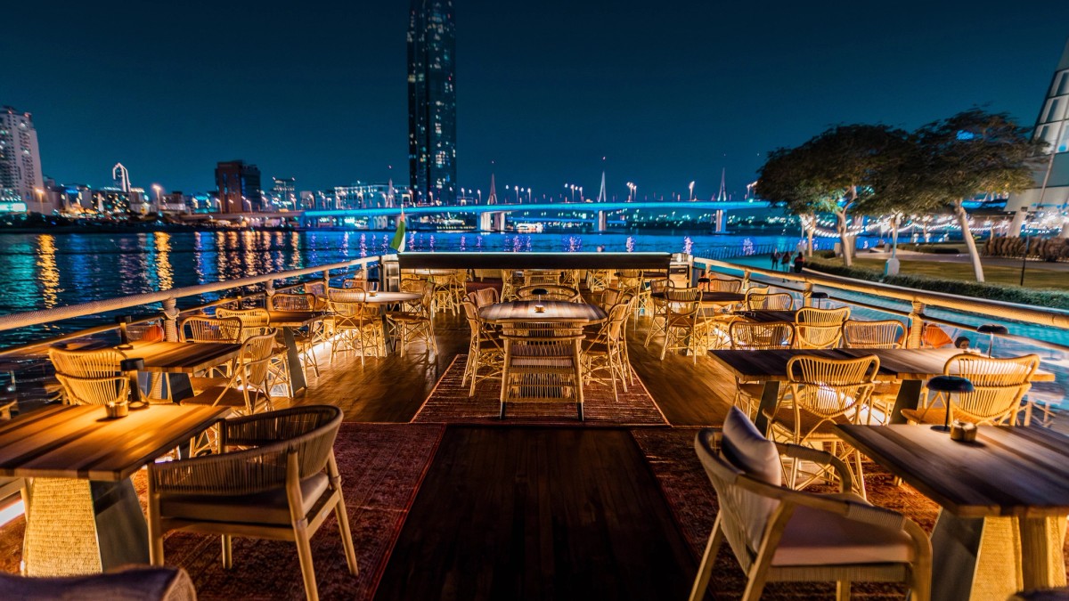 Tables set for dinner on the deck of a dhow, the Dubai skyline lit at night