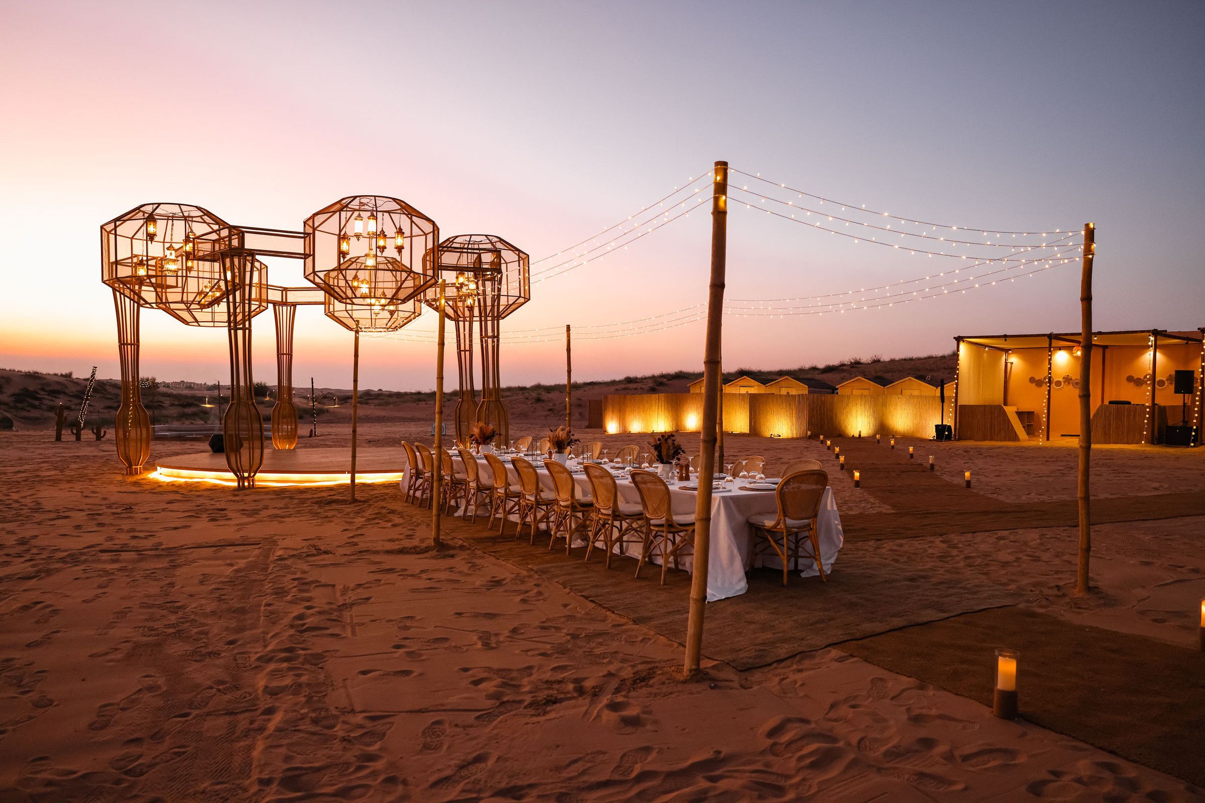 A long dinner table under lantern sculptures at a desert camp at dusk