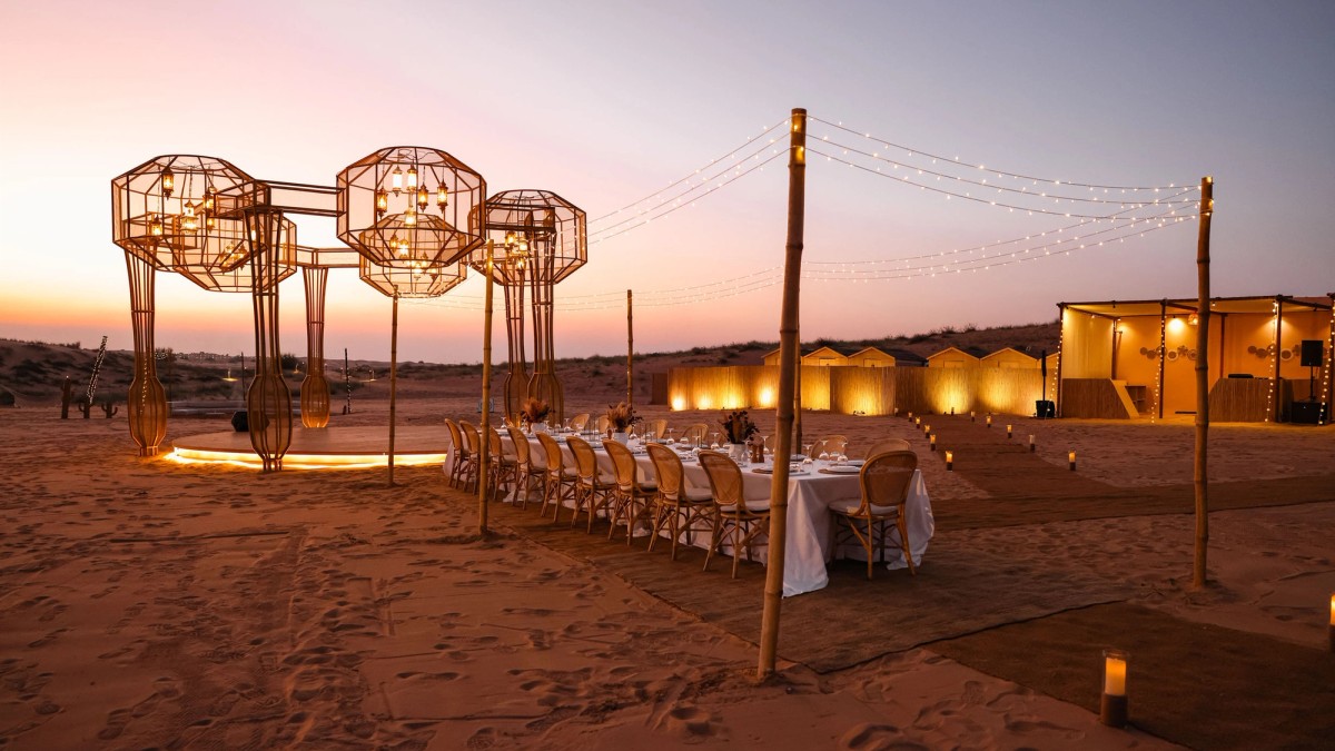 A long dinner table under lantern sculptures at a desert camp at dusk