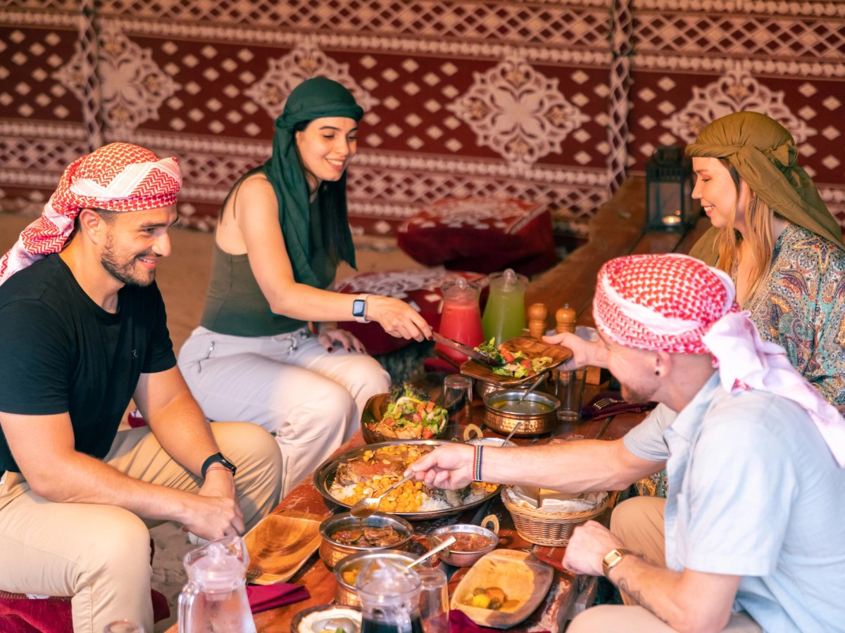 Guests in keffiyehs sharing a traditional dinner with their host in a desert camp majlis