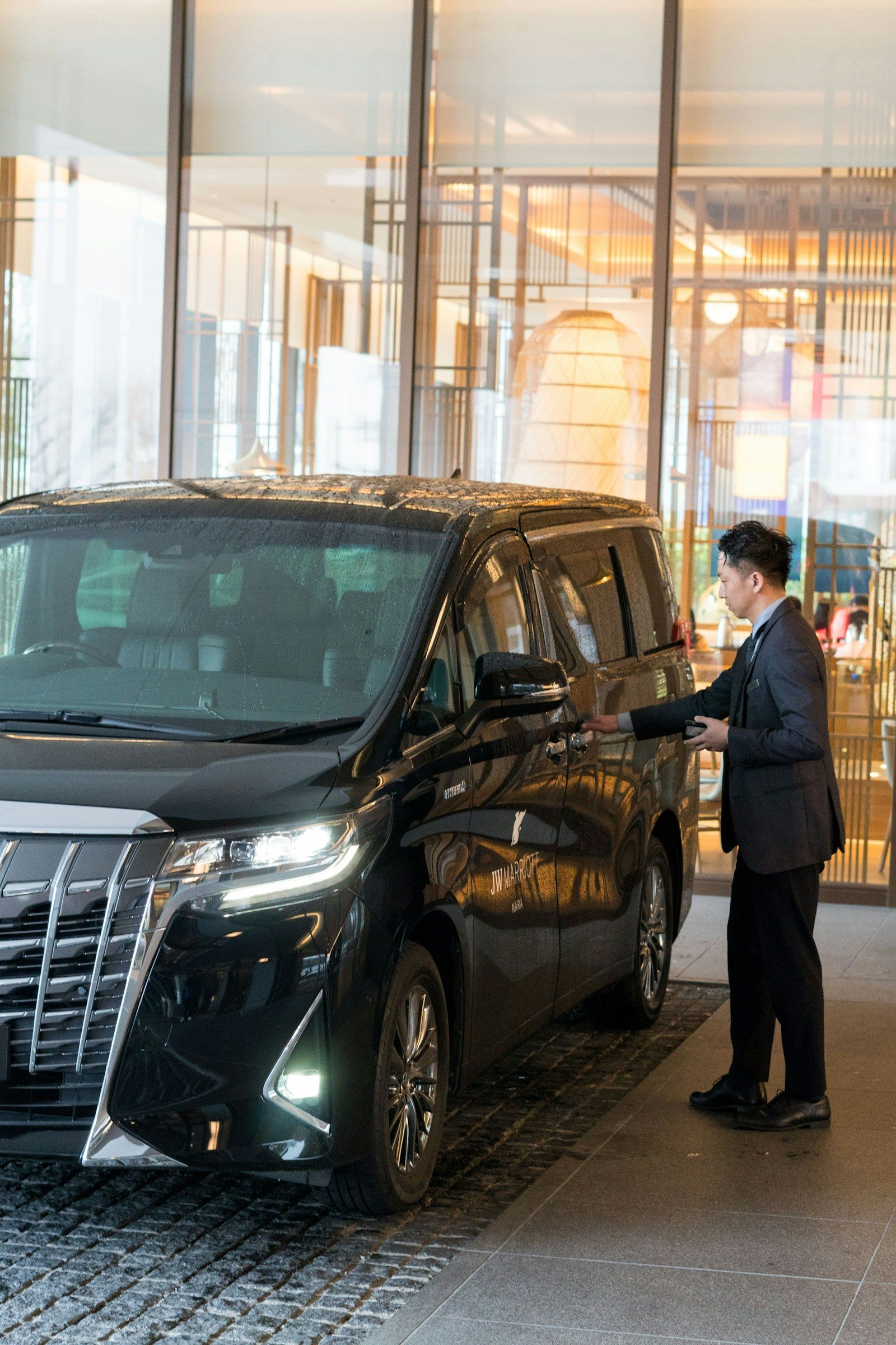 A chauffeur in a suit opening the door of a black people-carrier at a hotel entrance