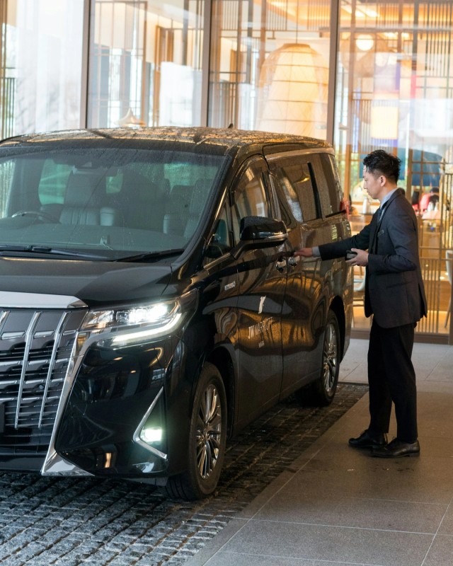 A chauffeur in a suit opening the door of a black people-carrier at a hotel entrance