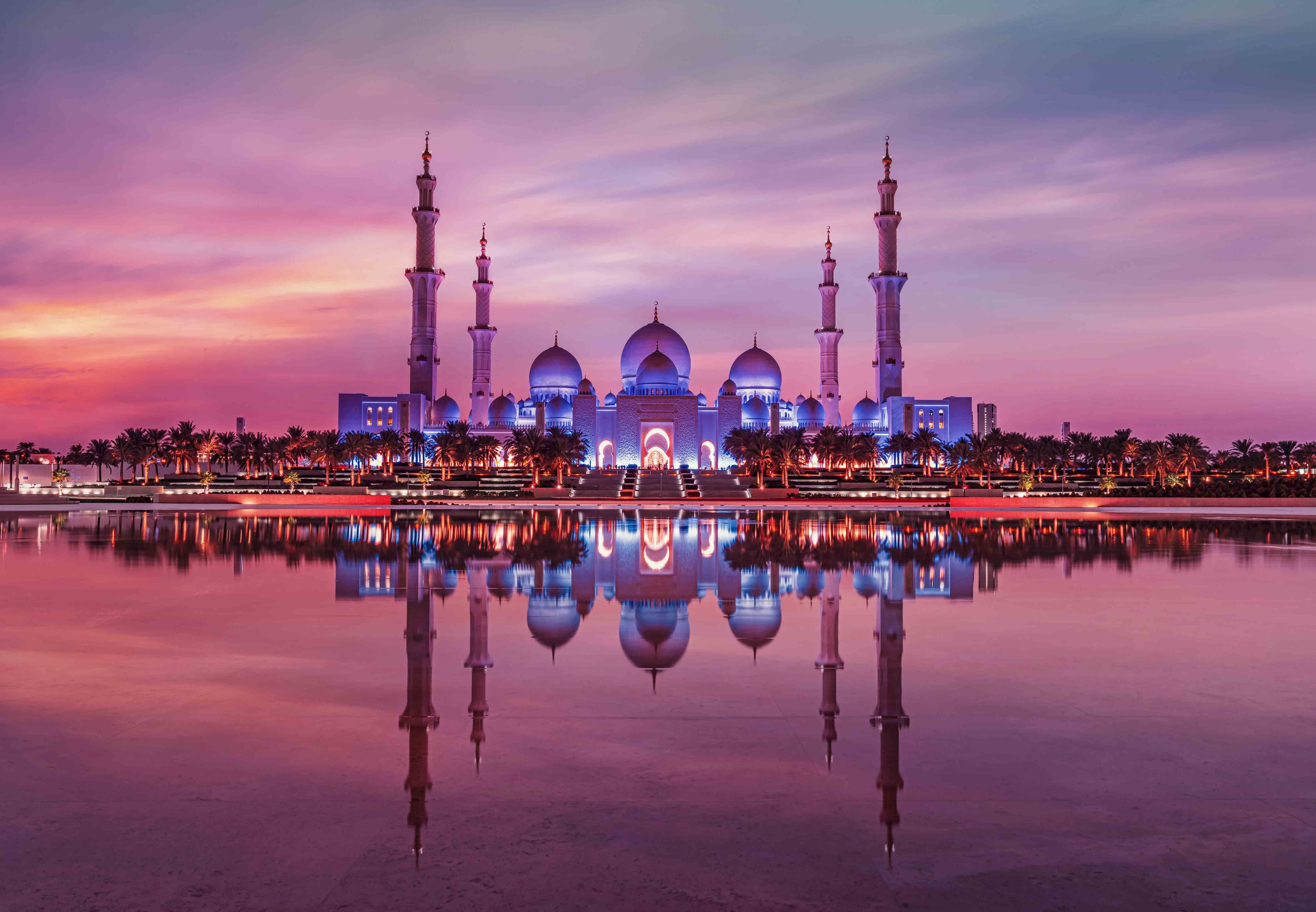 The mosque's minarets and domes glowing violet, mirrored in the reflecting pools