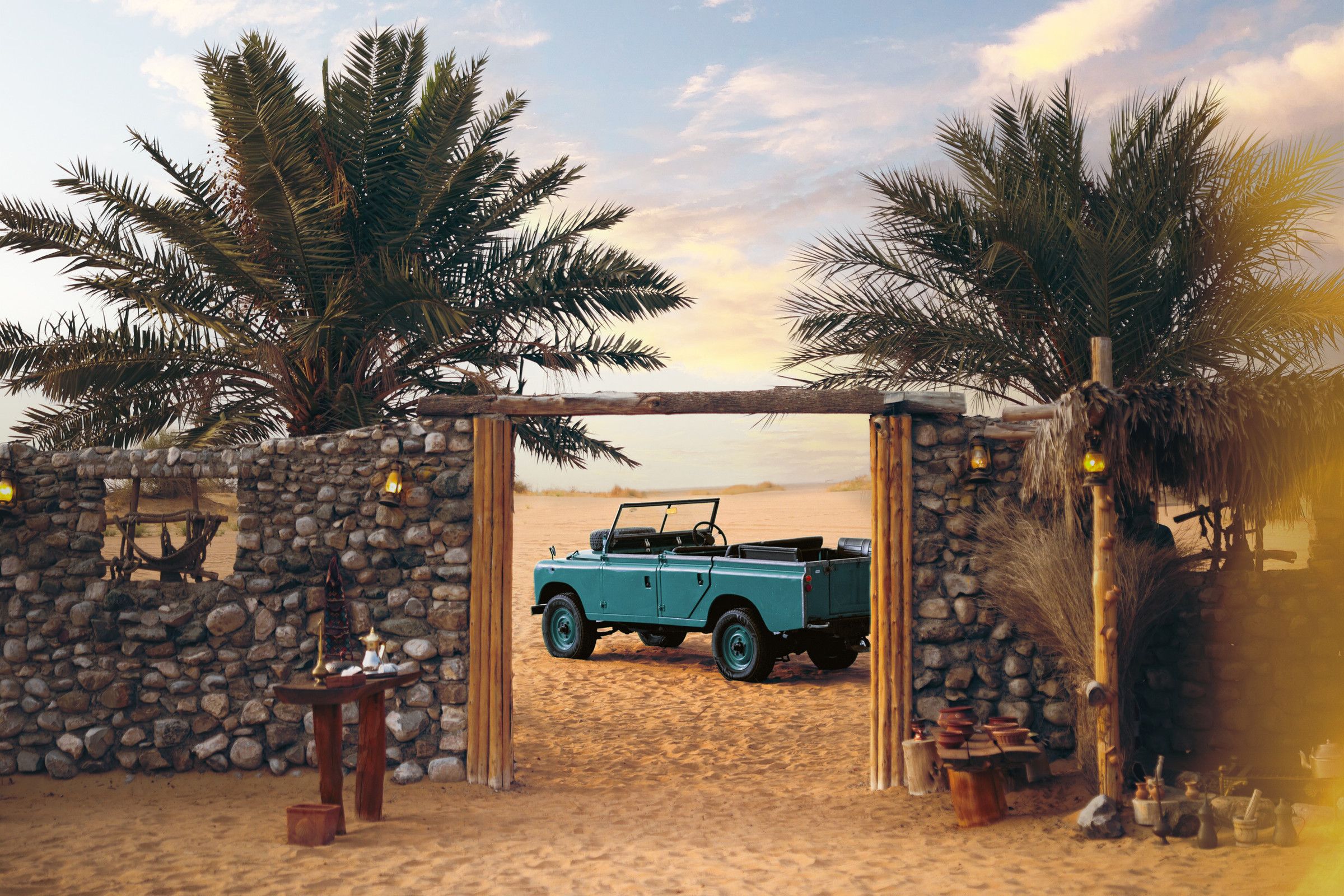 A vintage Land Rover framed by a palm-shaded stone gateway in the desert