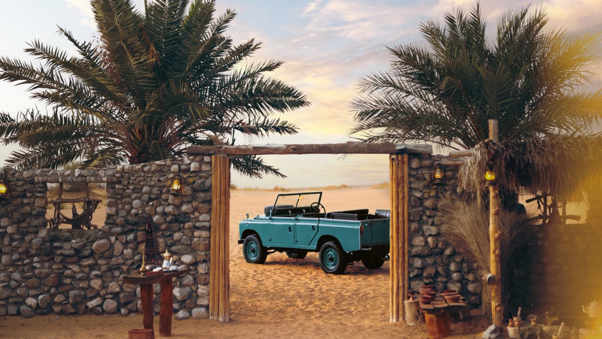 A vintage Land Rover framed by a palm-shaded stone gateway in the desert