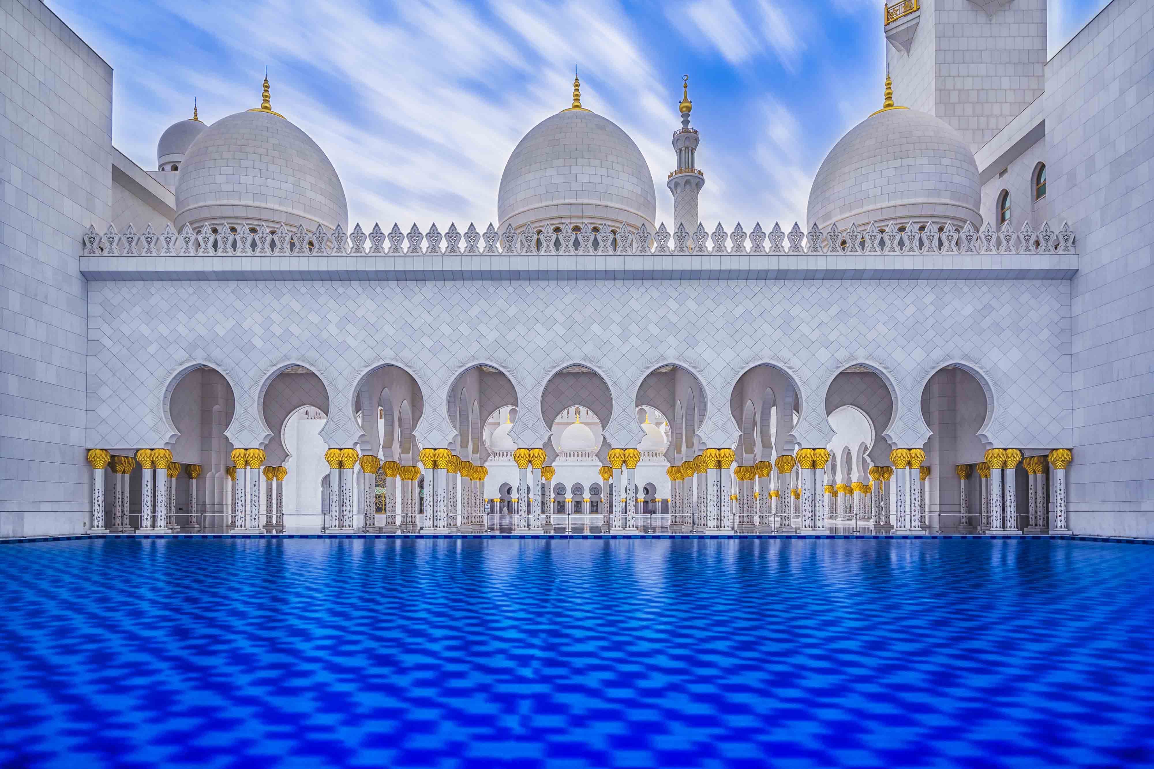 The white marble courtyard arcade and domes of the Sheikh Zayed Grand Mosque, Abu Dhabi, mirrored in a blue reflecting pool
