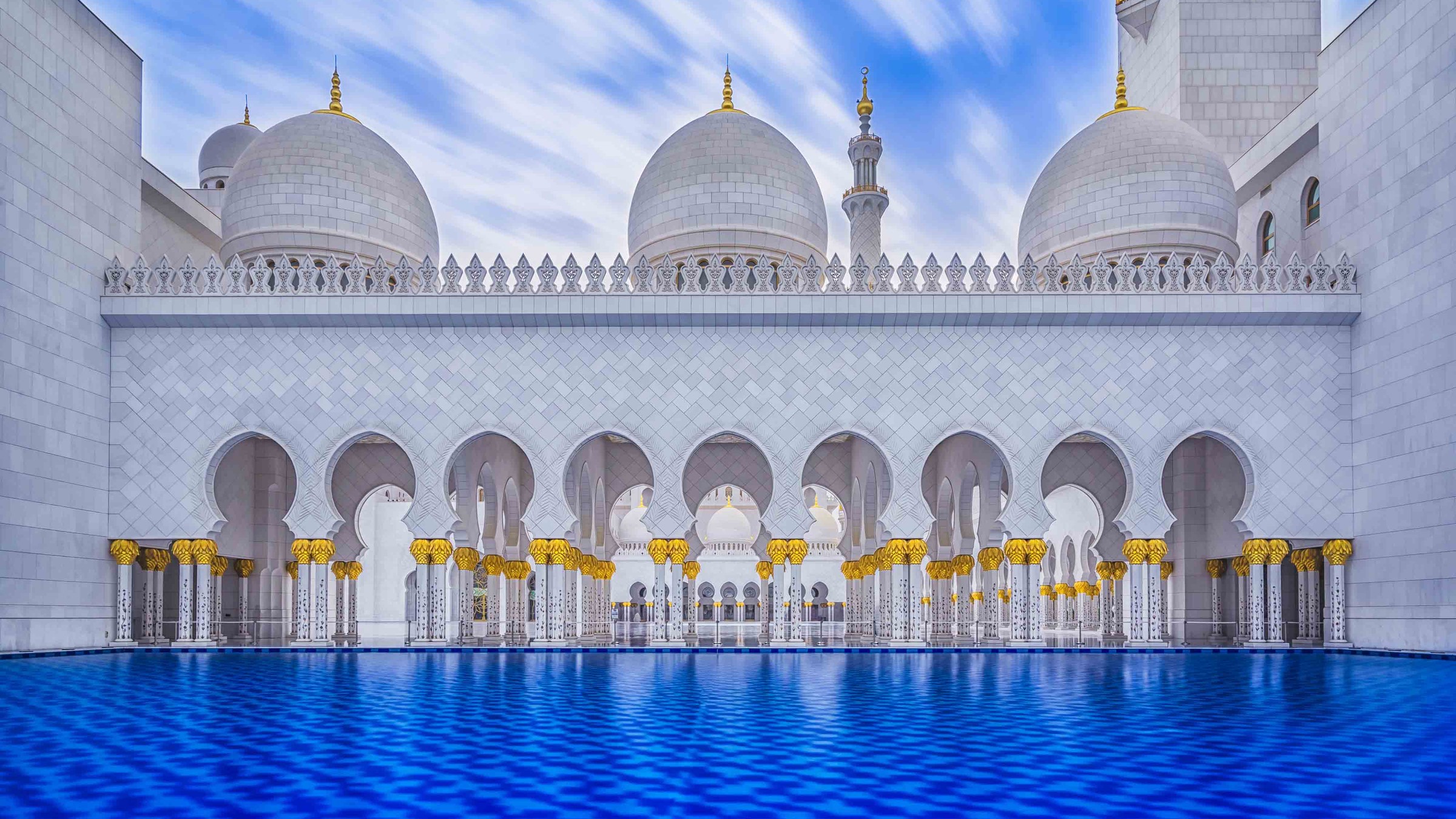 The white marble courtyard arcade and domes of the Sheikh Zayed Grand Mosque, Abu Dhabi, mirrored in a blue reflecting pool