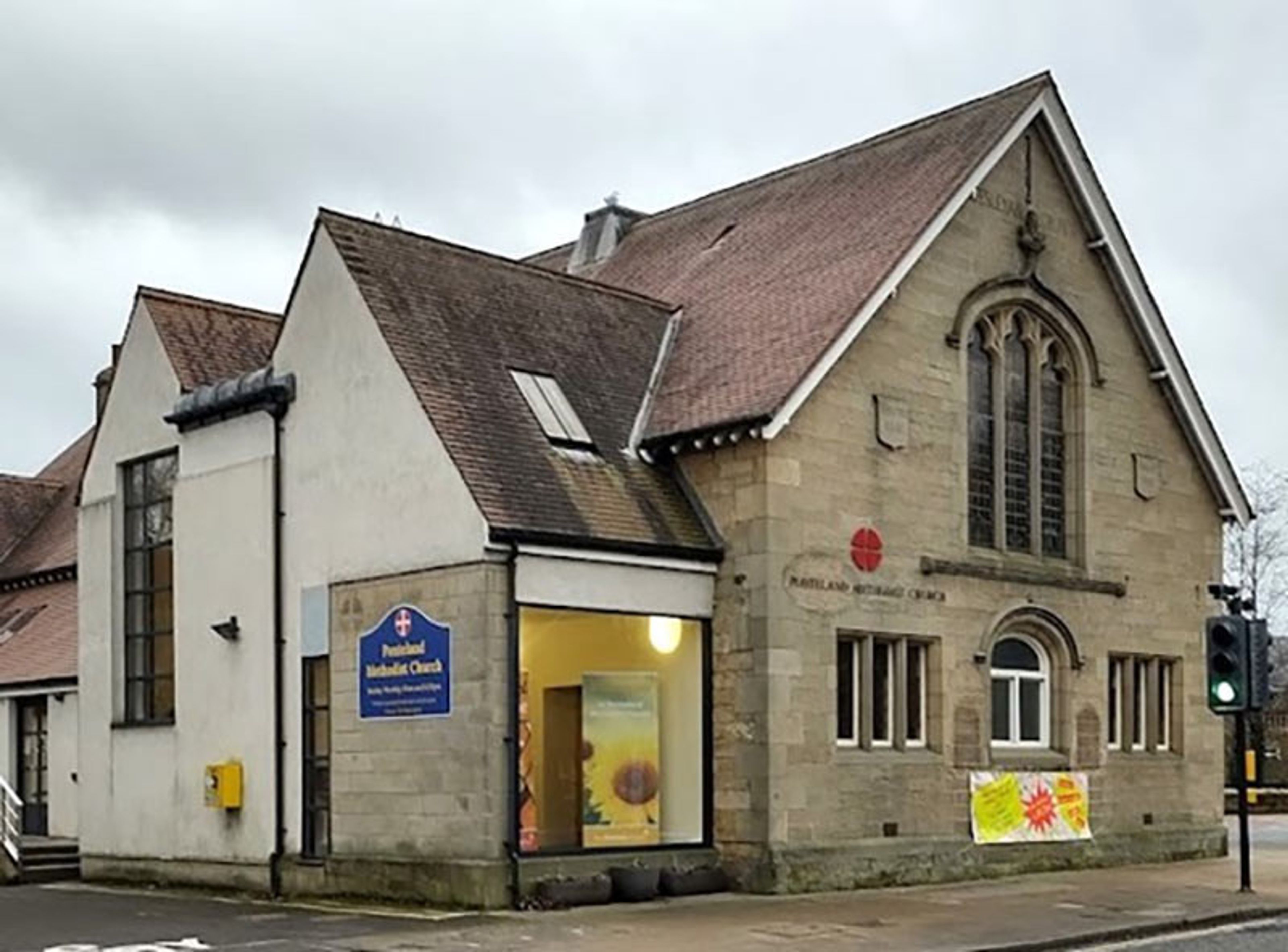 A stone Methodist church with a large traceried window and a modern rendered extension, on a street corner