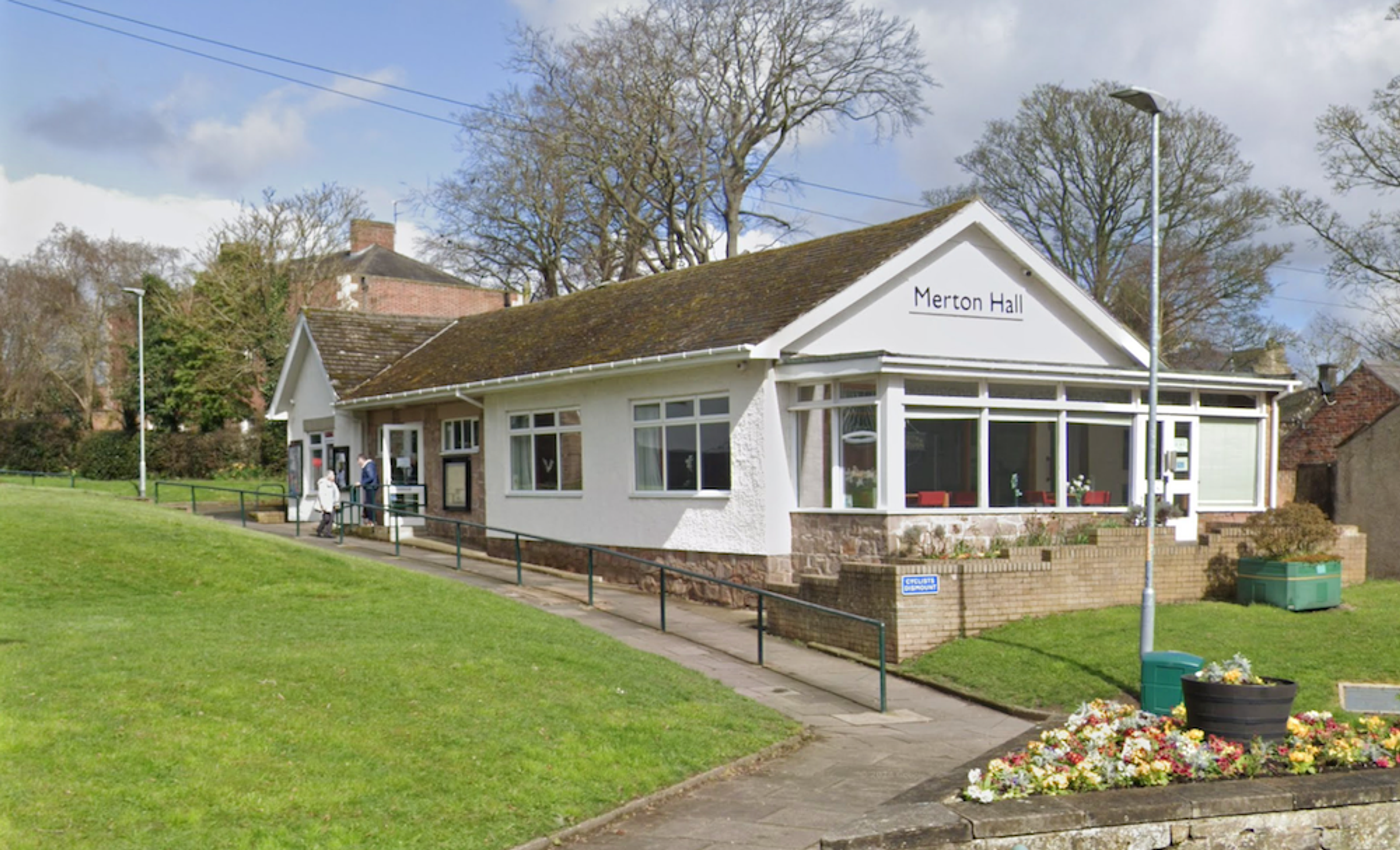 A white-painted single-storey community hall with a sign reading Merton Hall, a ramped path and flower beds in front