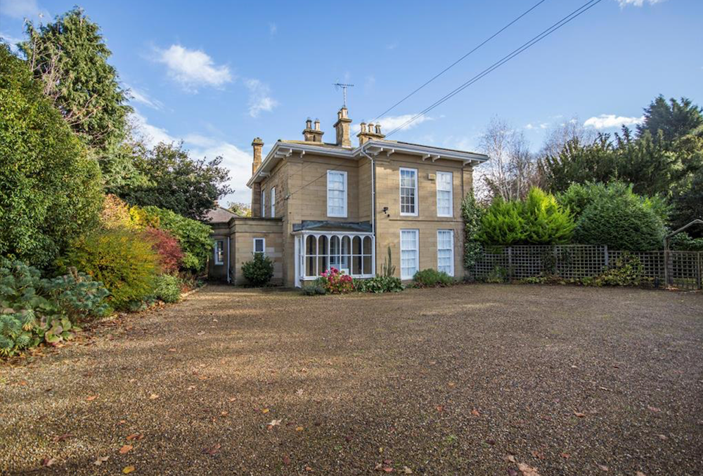 A stone Victorian villa with tall chimneys and a bay window, set behind a gravel drive and mature shrubs