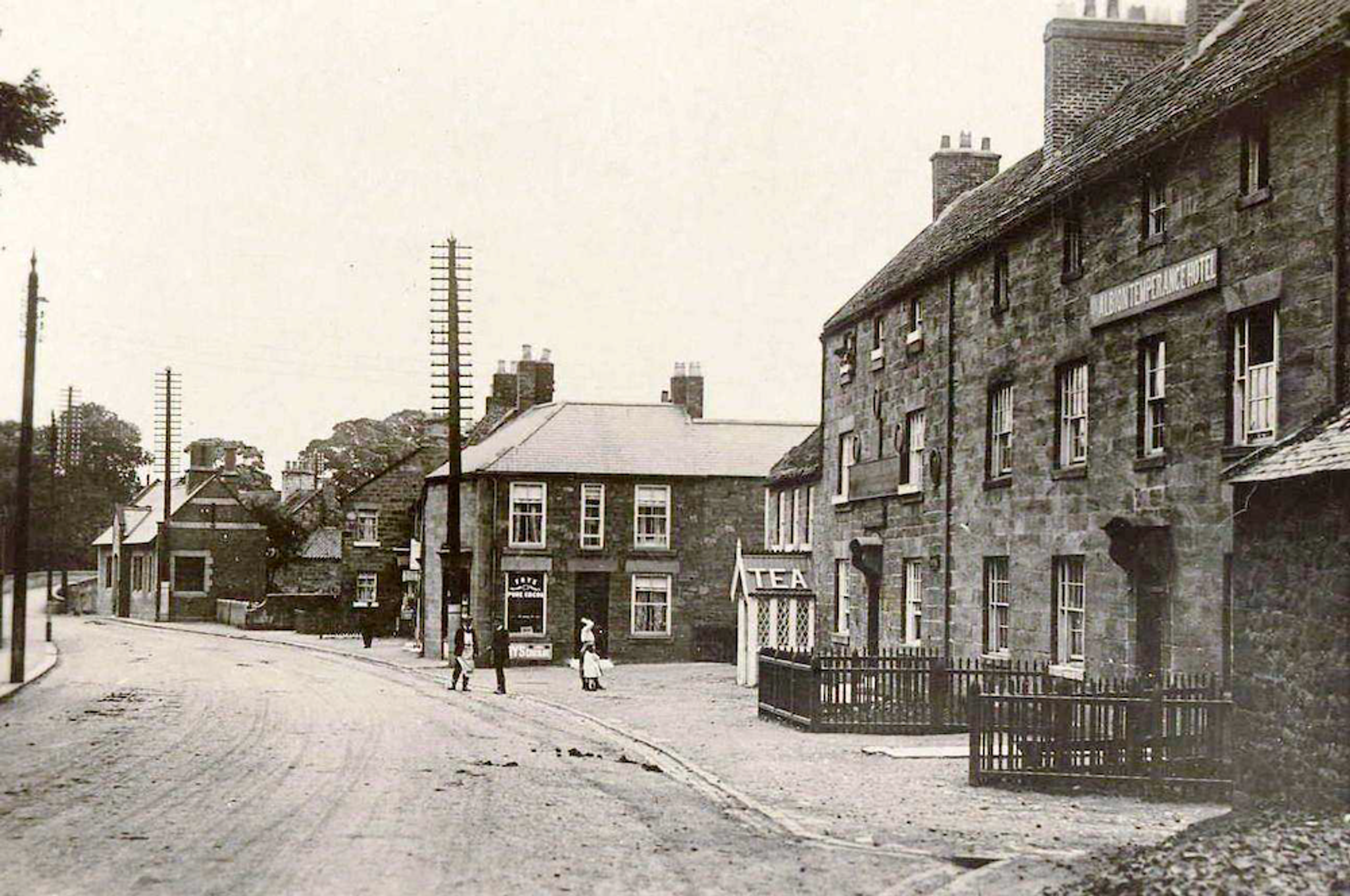 Black and white photograph of a village street lined with stone buildings, a shop with a TEA sign, telegraph poles and people standing in the road