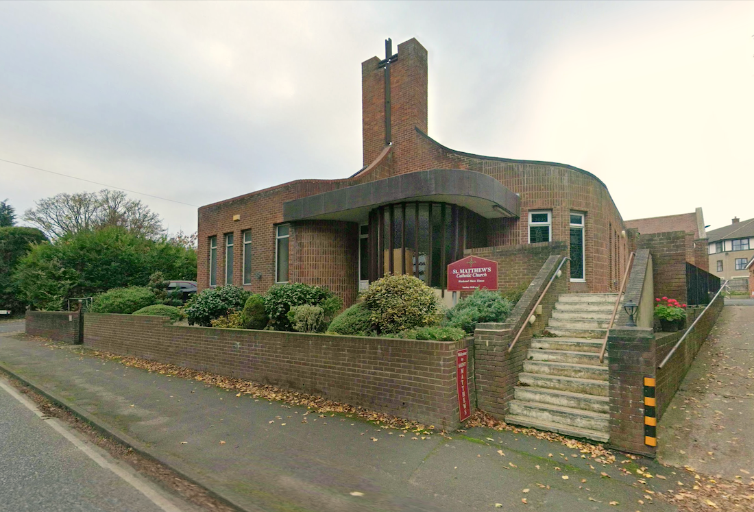 A modern brick Roman Catholic church with a tall square tower and a canopied entrance, steps rising from the roadside