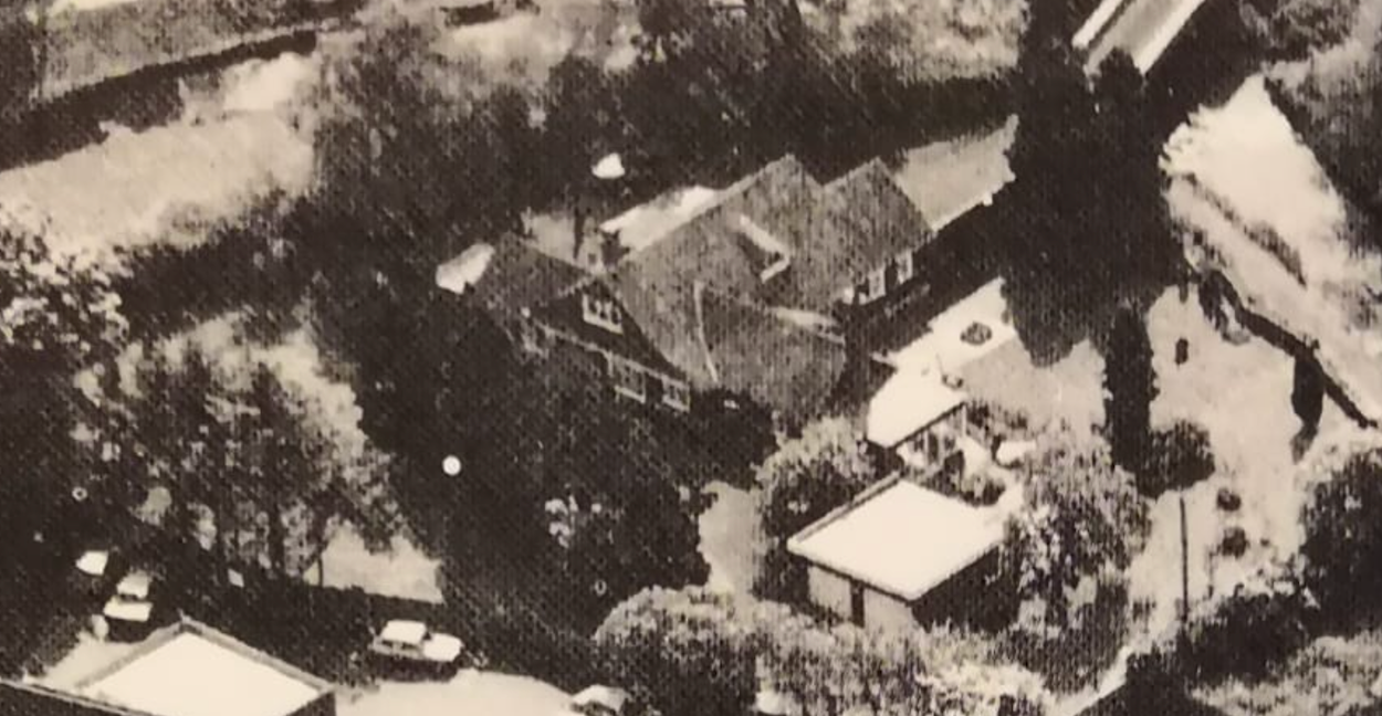 Grainy black and white aerial photograph of a large house with gabled roofs set among dense trees and gardens