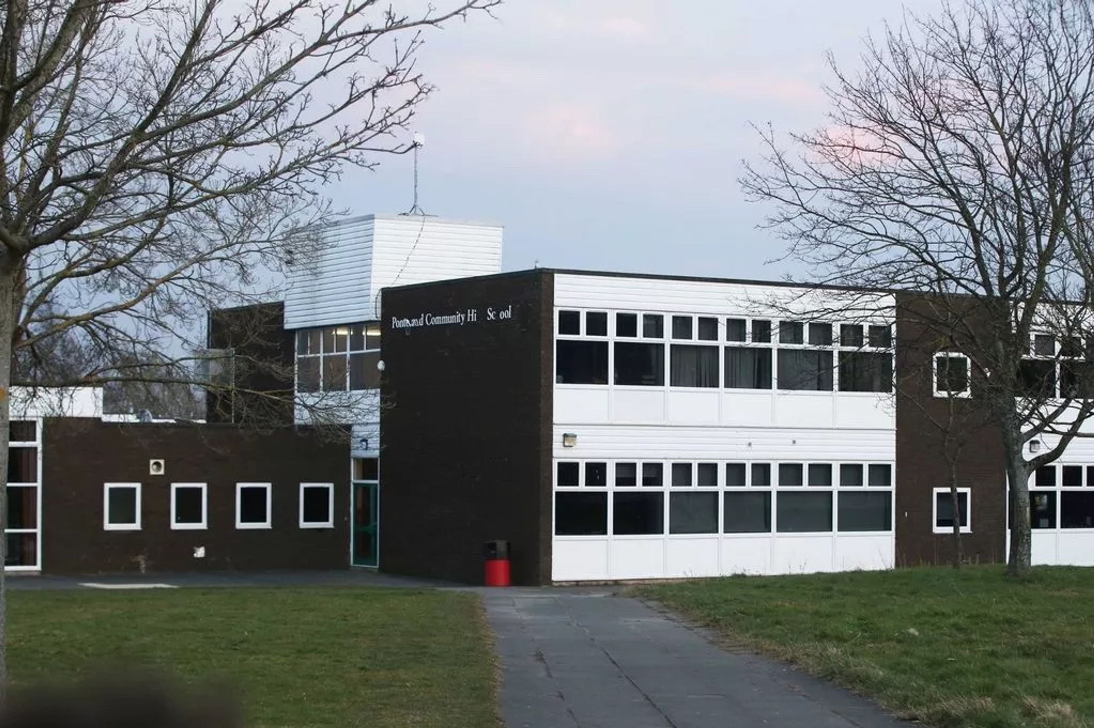 A 1960s two-storey school building in dark brick with long window bands and a rooftop clock tower, seen across the grass
