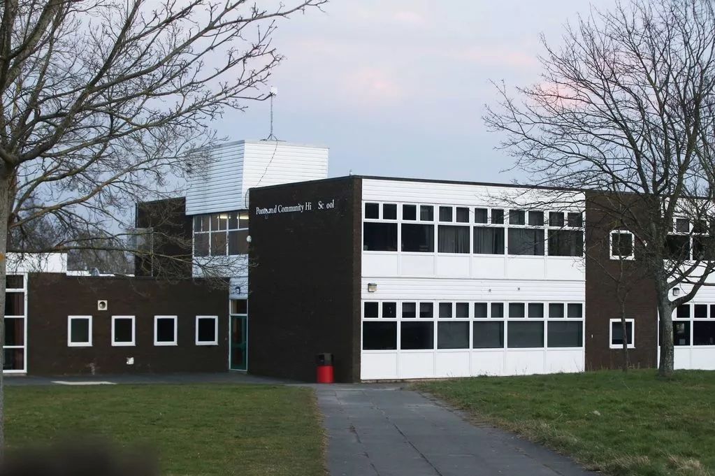 A 1960s two-storey school building in dark brick with long window bands and a rooftop clock tower, seen across the grass