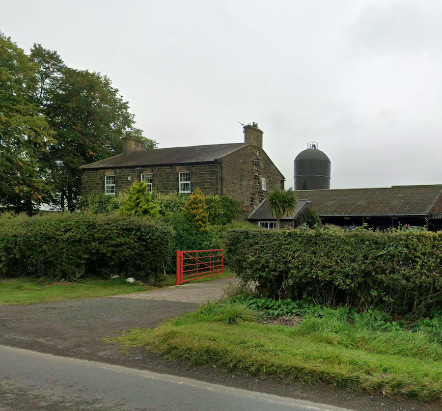 A stone farmhouse behind a hedge with a red metal field gate, farm buildings and a grain silo to the side
