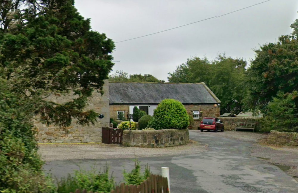 A long single-storey stone farm building with a slate roof and clipped hedge, set behind a stone wall at a road junction
