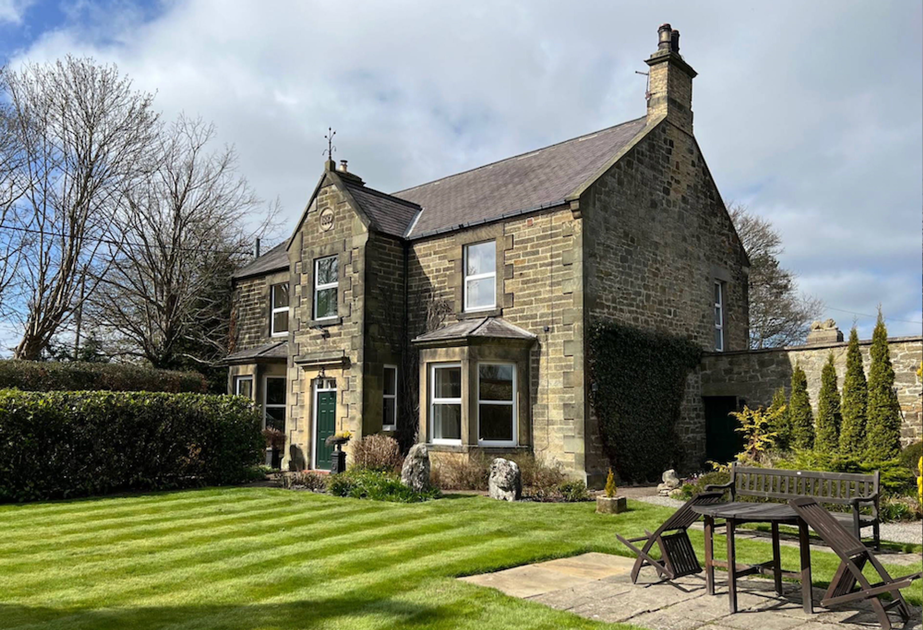A stone farmhouse with a bay window and gabled porch, standing behind a striped lawn with a garden table and bench