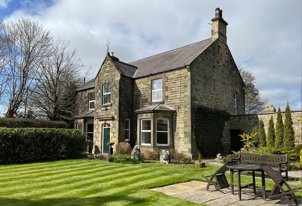 A stone farmhouse with a bay window and gabled porch, standing behind a striped lawn with a garden table and bench