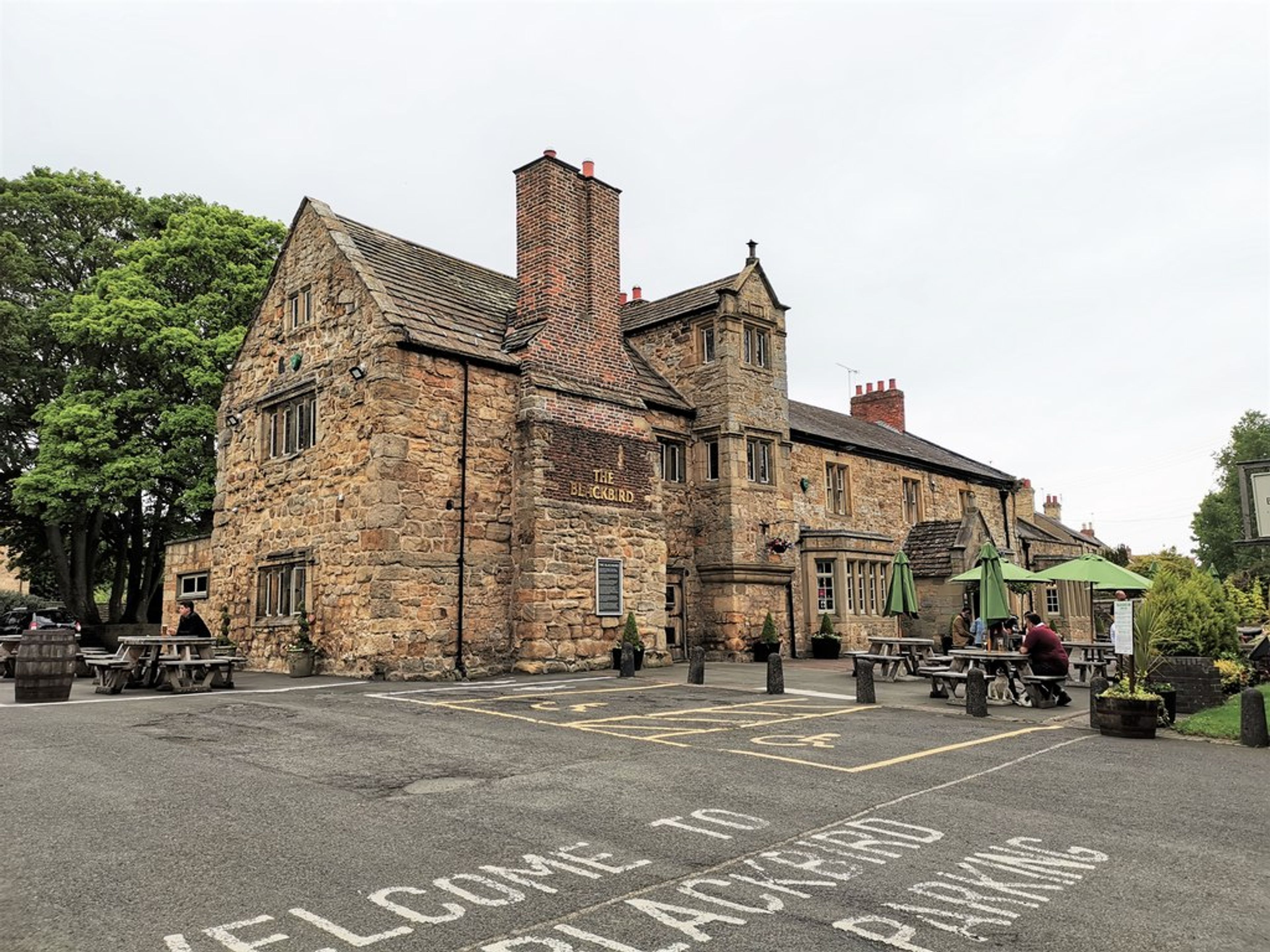 A stone pub with a tall chimney breast and mullioned windows, picnic benches and parasols on the car park in front