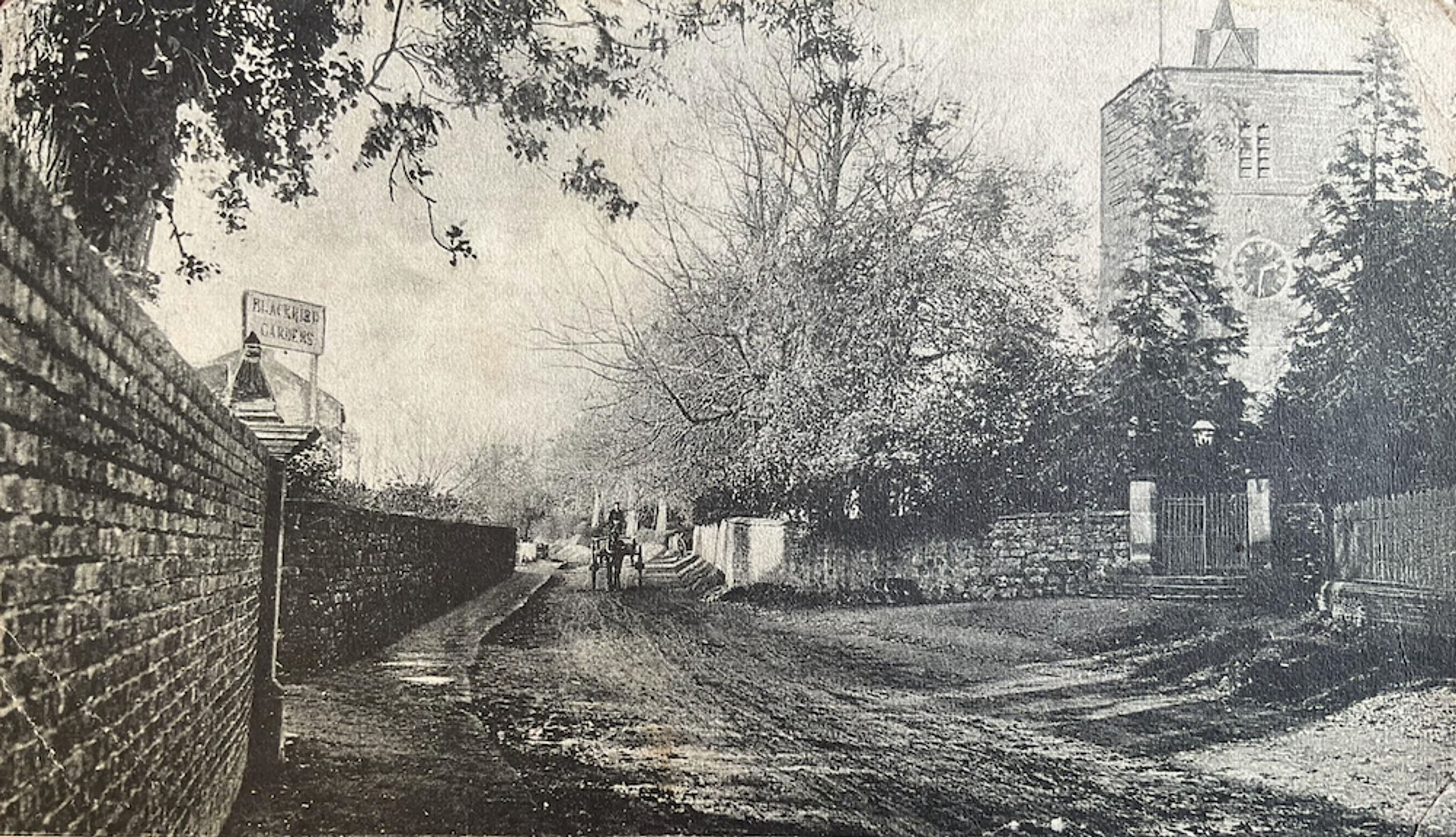 Old black and white photograph of an unmade lane running between a brick wall and a stone wall, a church tower rising through the trees