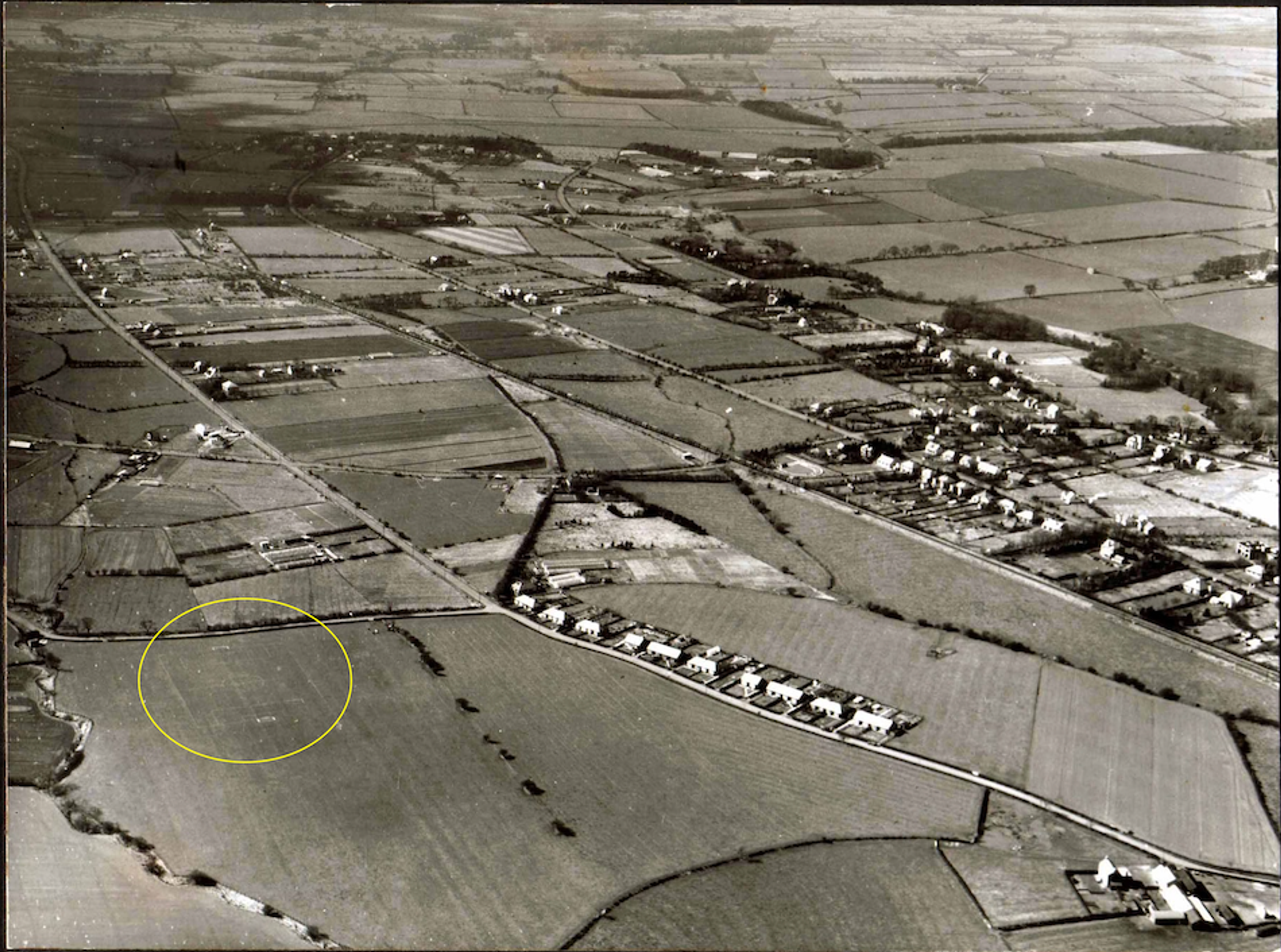 Black and white aerial photograph of farmland and a village, with a yellow ellipse marking an open field beside a road