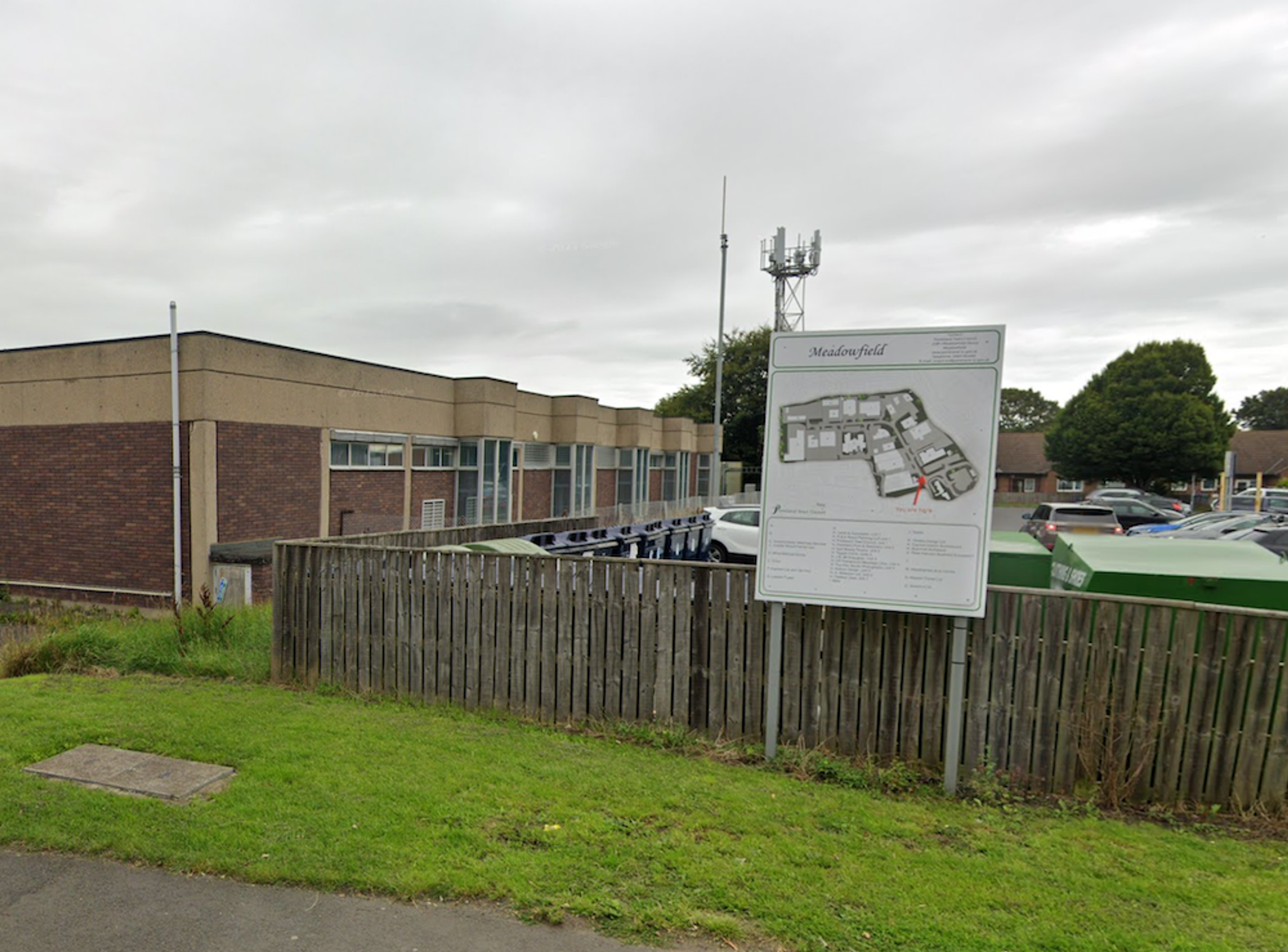 A low industrial unit behind a wooden fence, with a Meadowfield estate map board and a telecommunications mast alongside