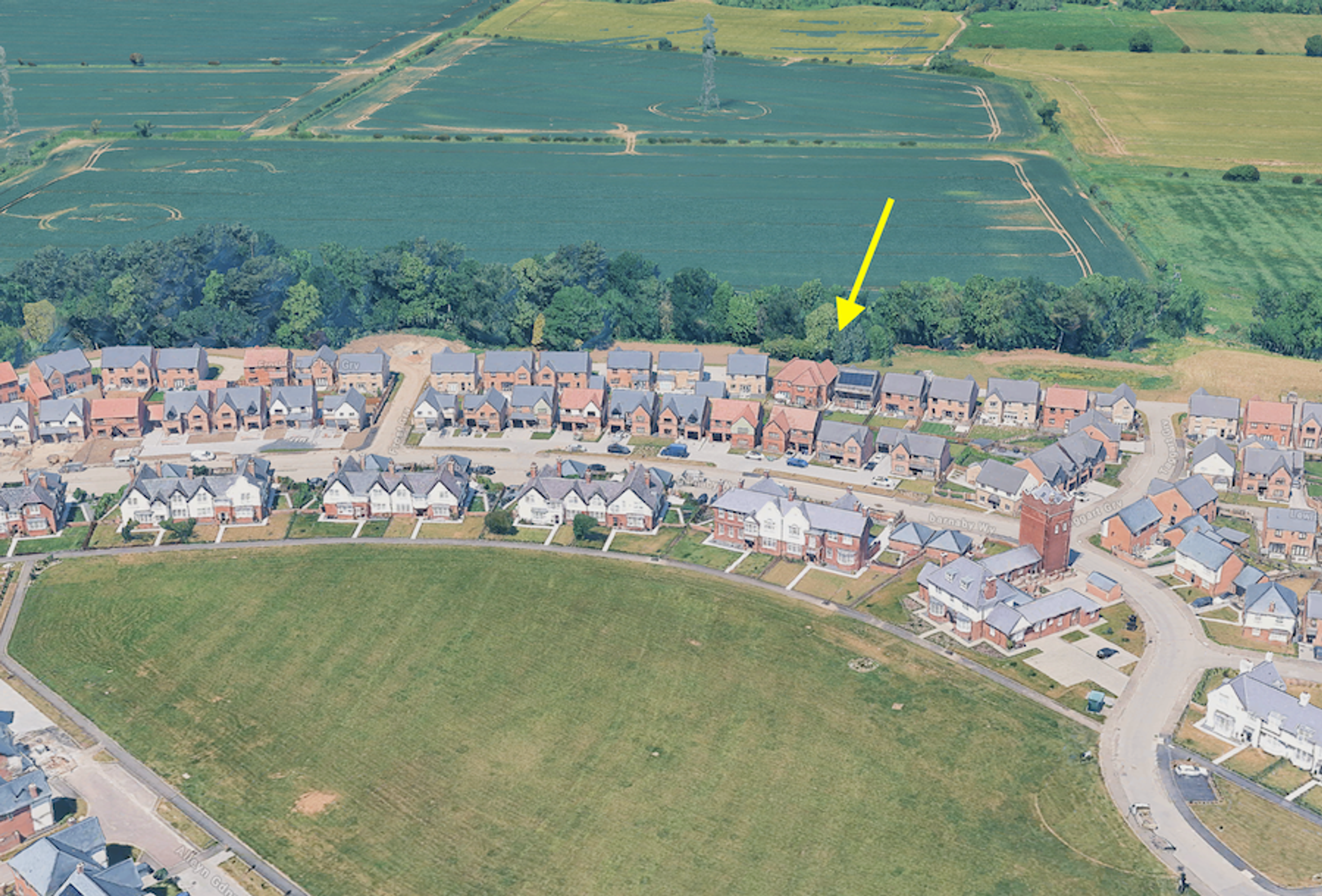 Aerial view of a modern housing estate beside open fields, a yellow arrow pointing to a spot at the edge of the trees