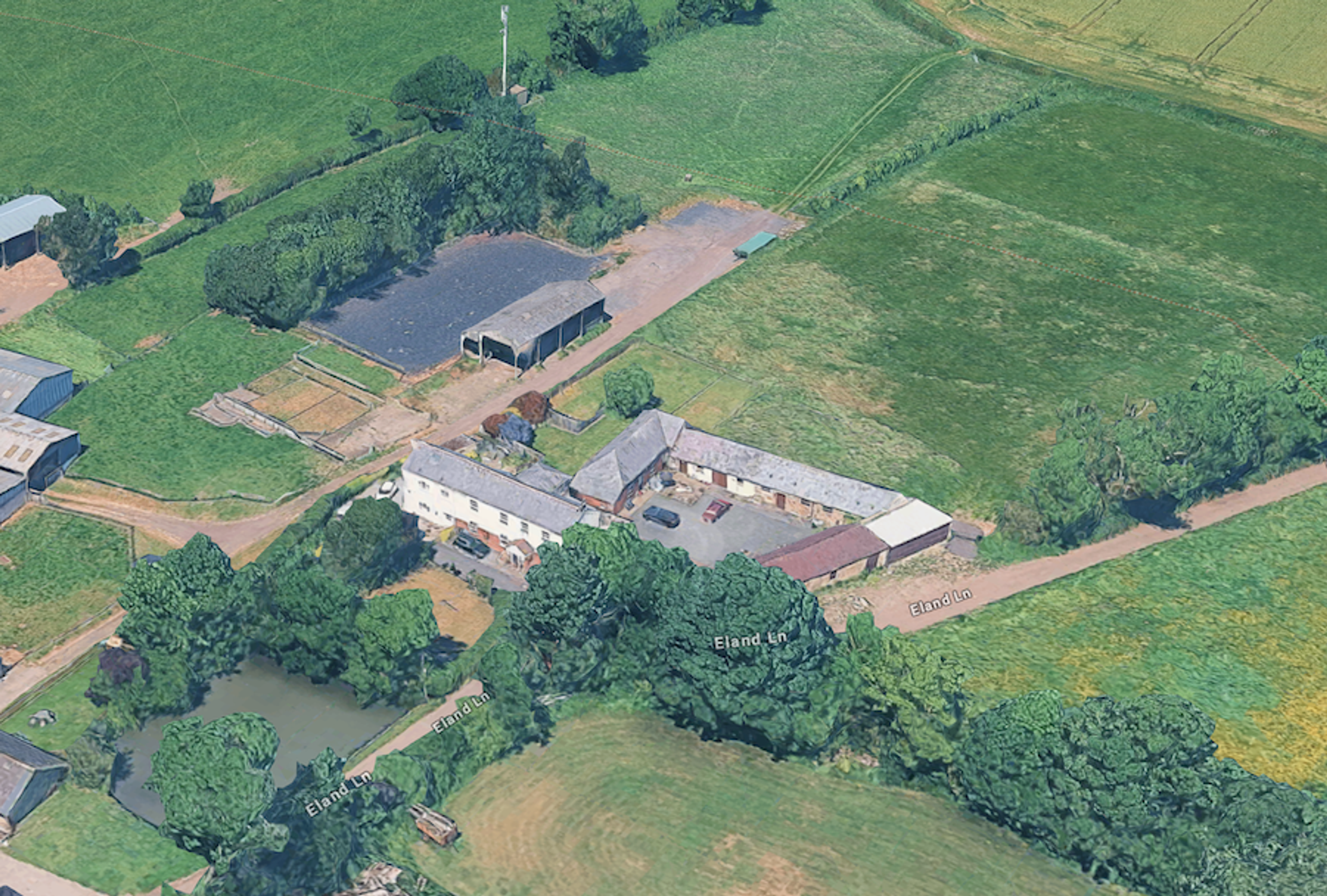Aerial view of a farmstead with a long range of outbuildings, a farmhouse and a pond, surrounded by green fields