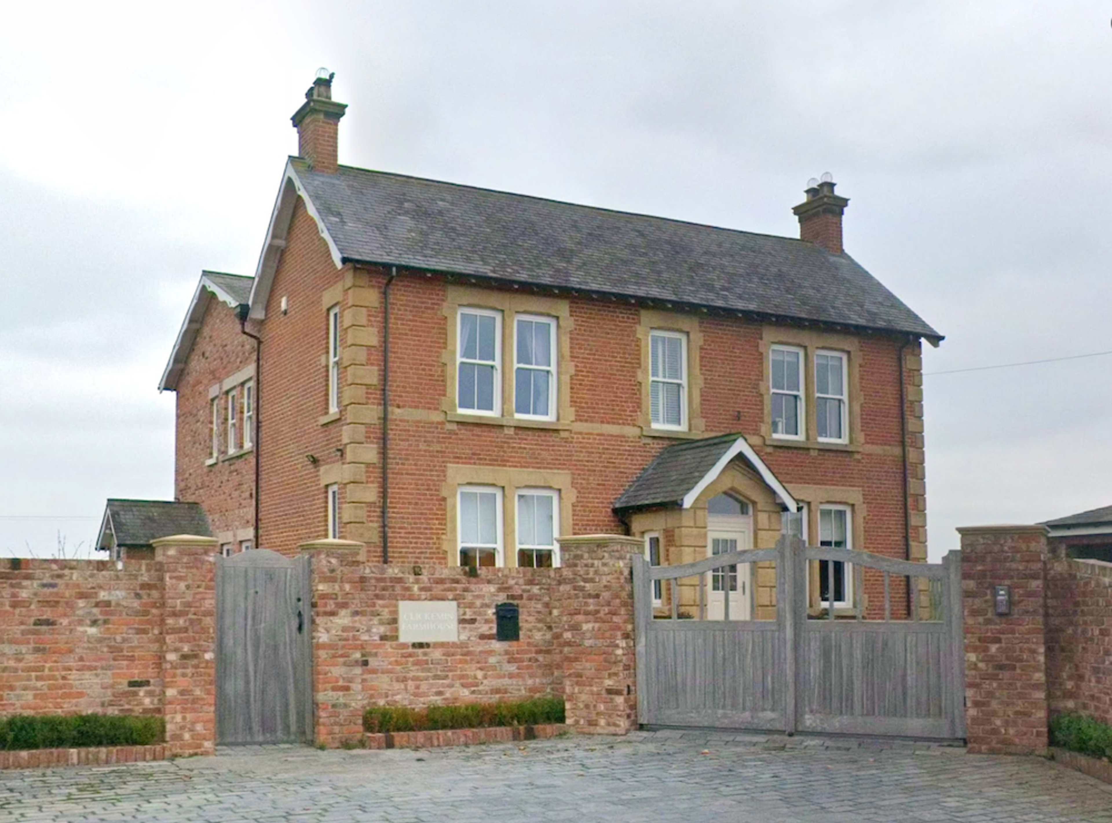 A converted brick and stone farmhouse with a gabled porch, behind a brick wall and timber gates on a cobbled forecourt