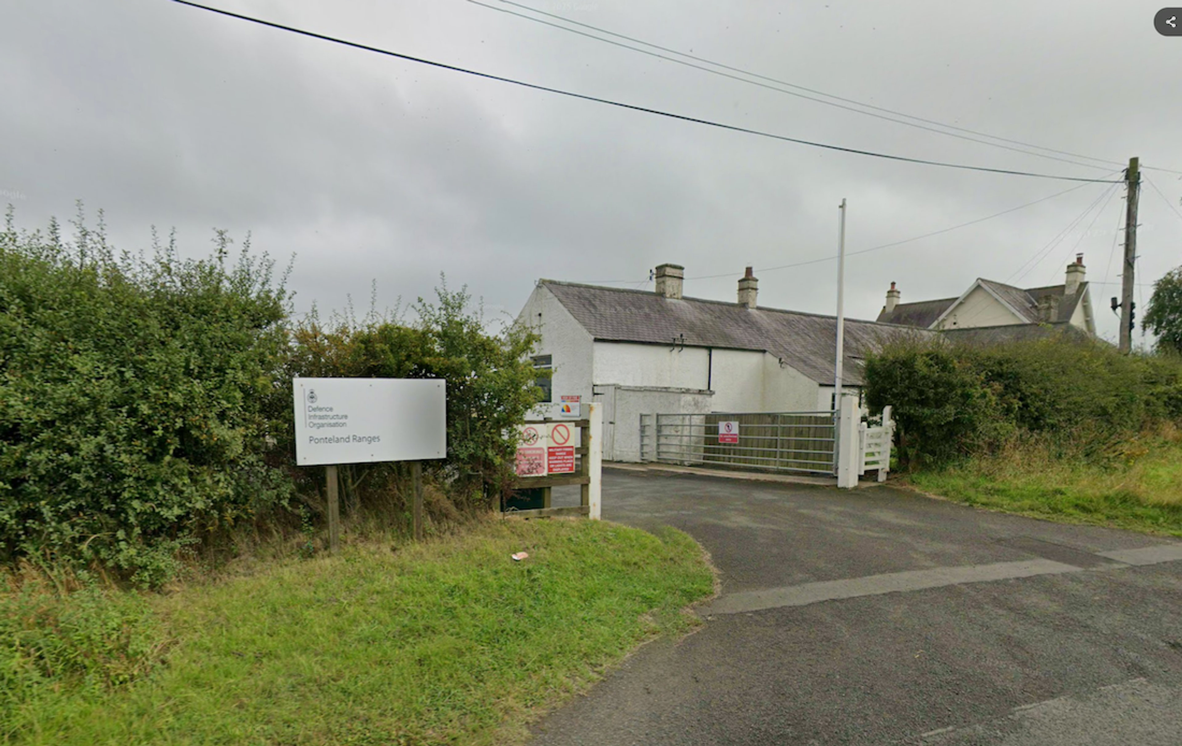 A gated entrance off a country road with an information sign and warning notices, whitewashed buildings beyond the hedge