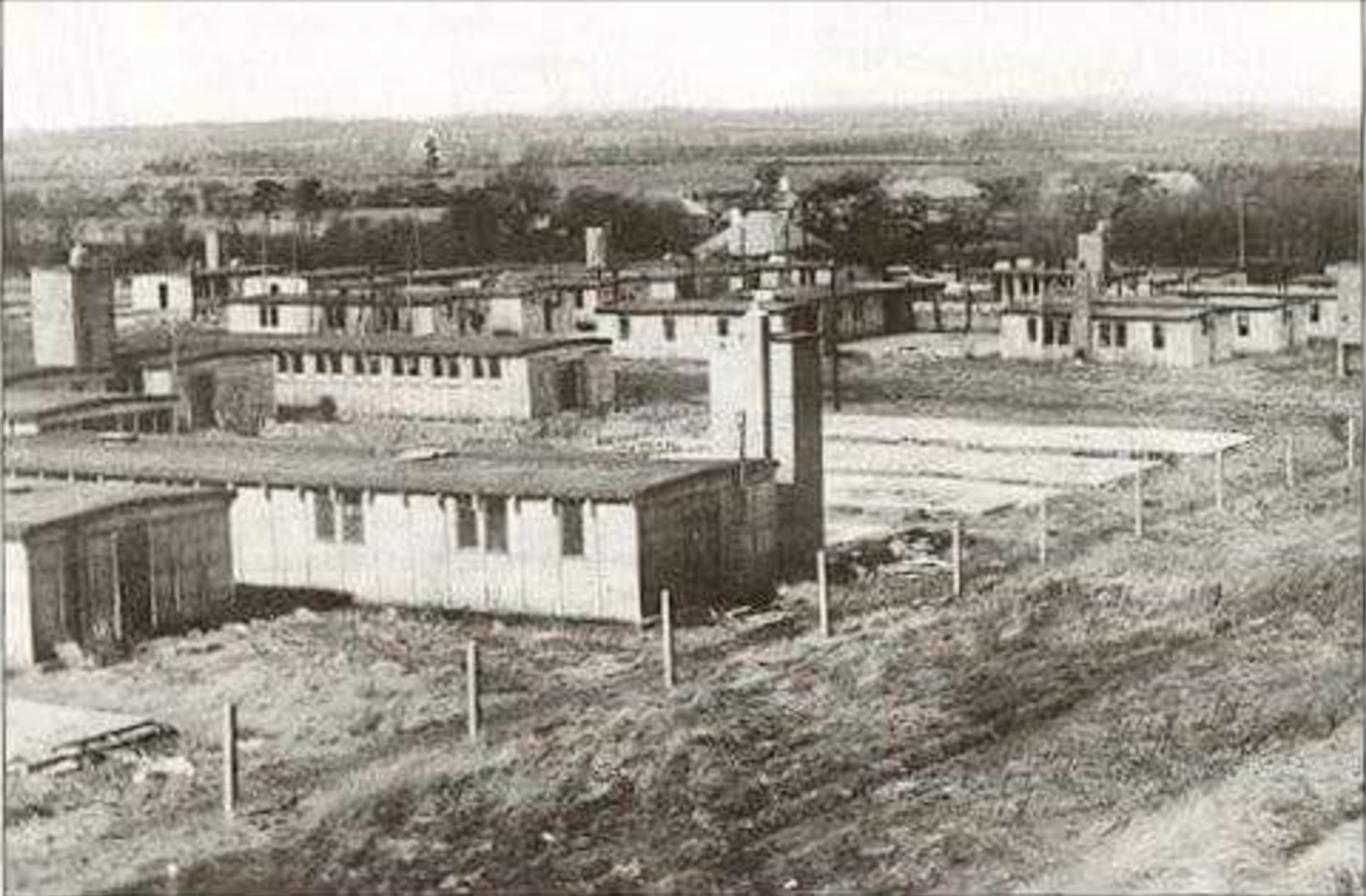 Black and white photograph of rows of single-storey prefabricated huts on open ground behind a wire fence