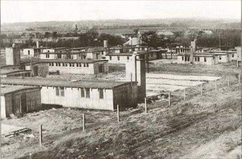 Black and white photograph of rows of single-storey prefabricated huts on open ground behind a wire fence
