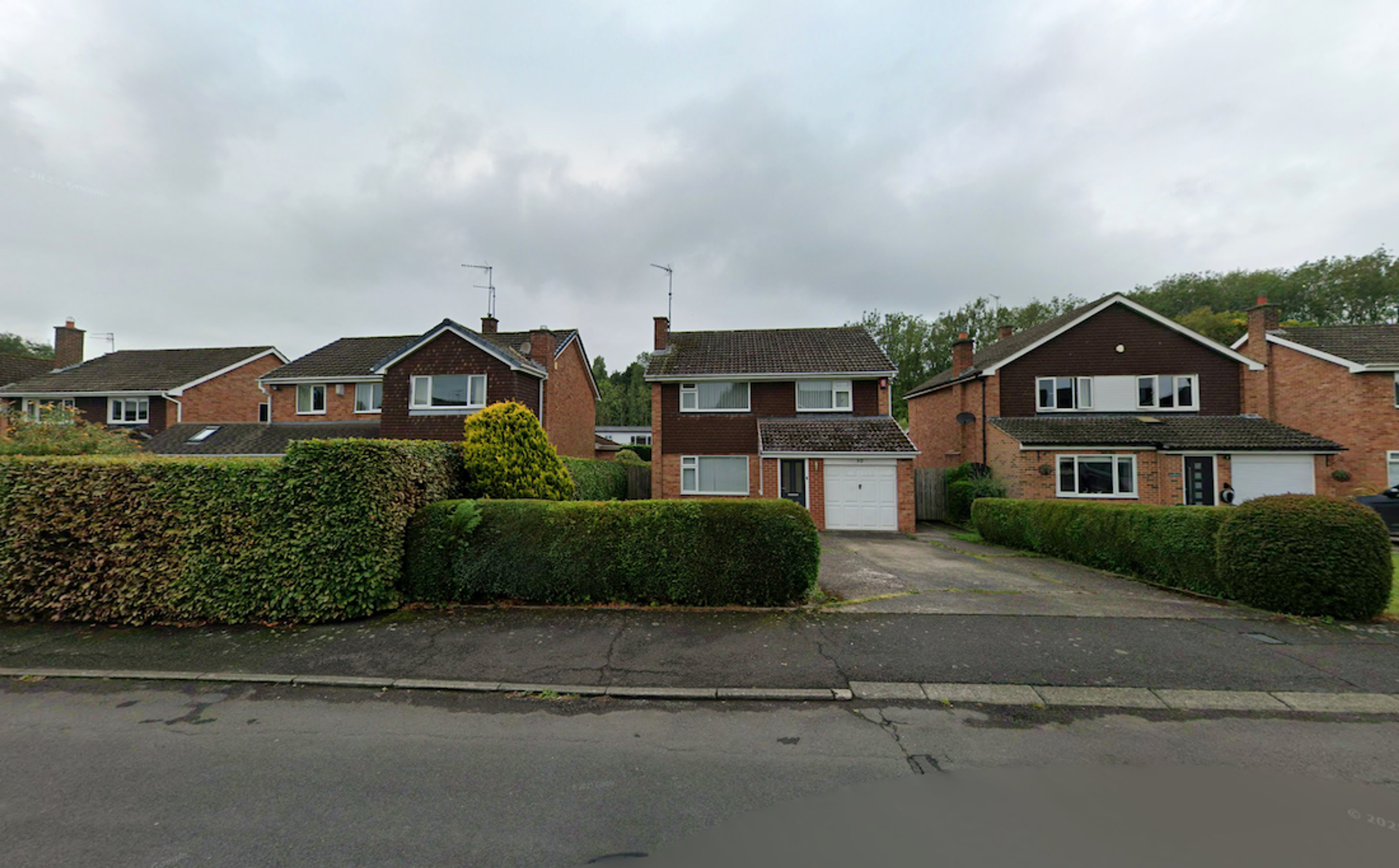 A row of 1960s detached brick houses with garages, set behind clipped hedges along a suburban street