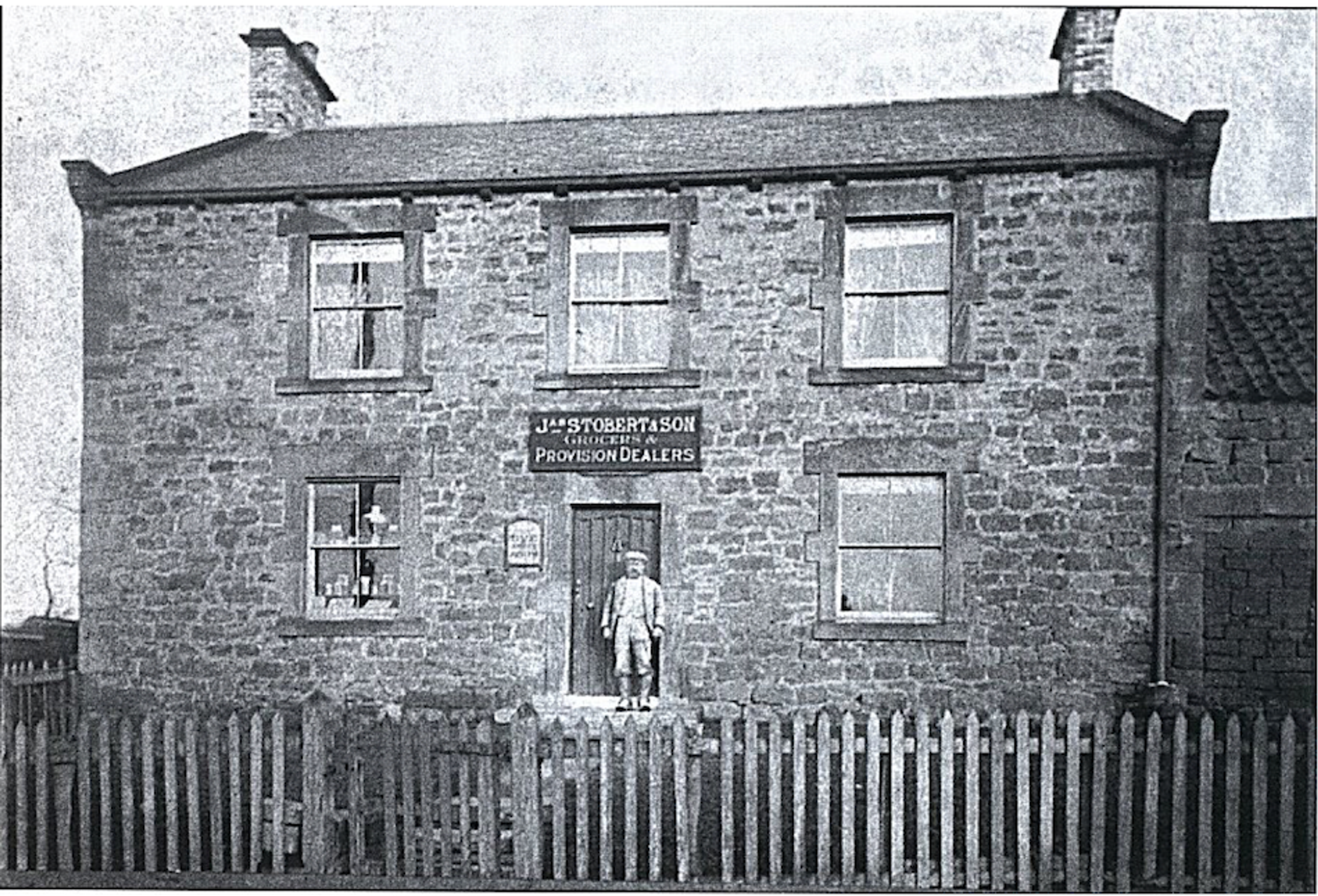 Black and white photograph of a two-storey stone shop and house with a sign reading Stobert and Son, Grocers and Provision Dealers, a man standing in the doorway