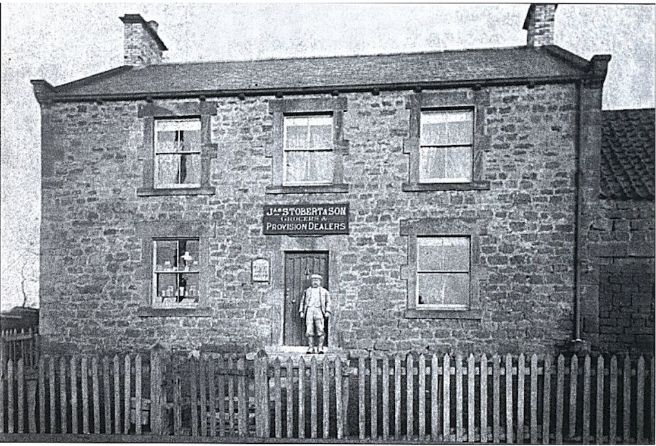 Black and white photograph of a two-storey stone shop and house with a sign reading Stobert and Son, Grocers and Provision Dealers, a man standing in the doorway