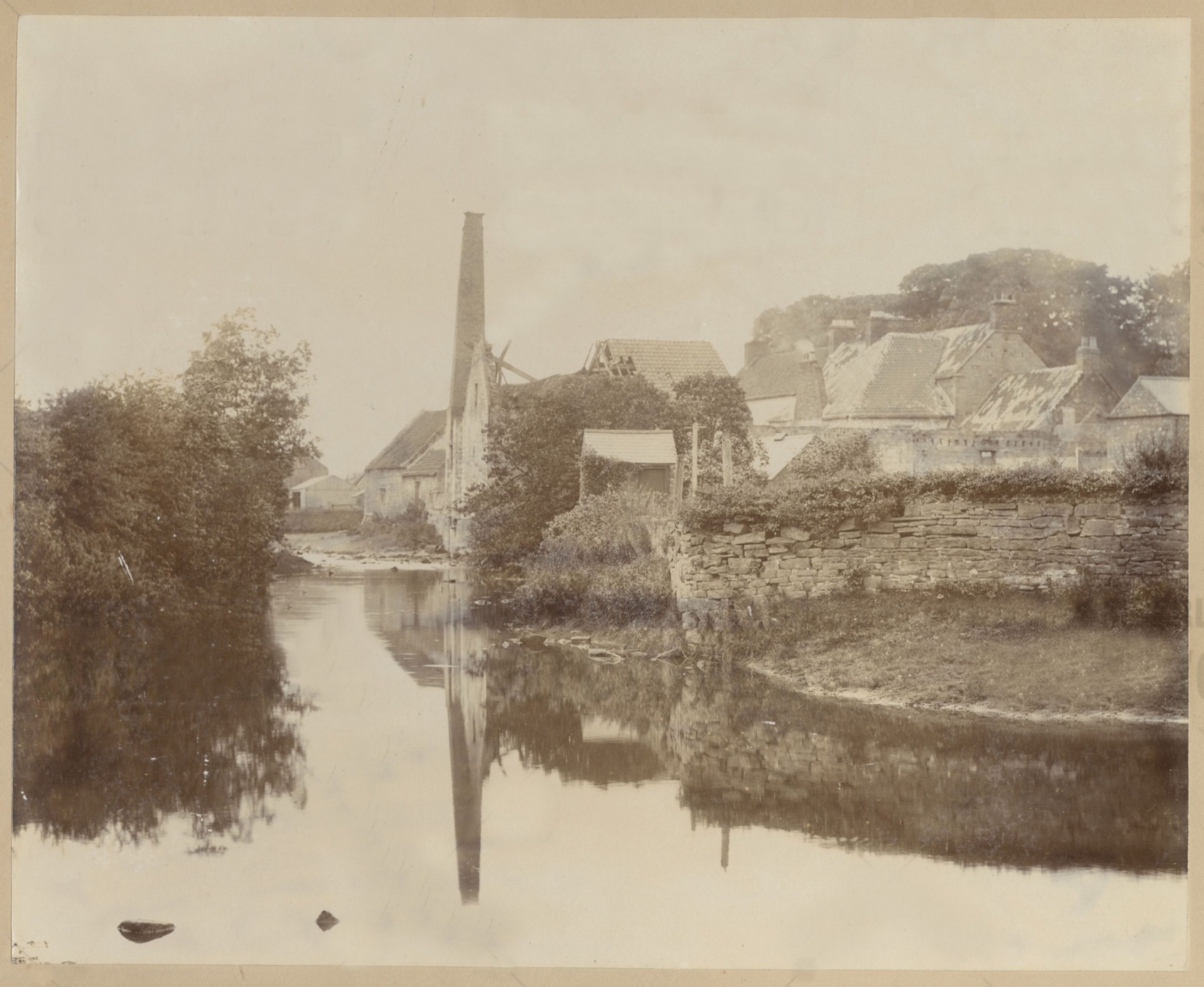 Sepia photograph of a river with buildings and rooftops rising behind a stone retaining wall, trees reflected in still water