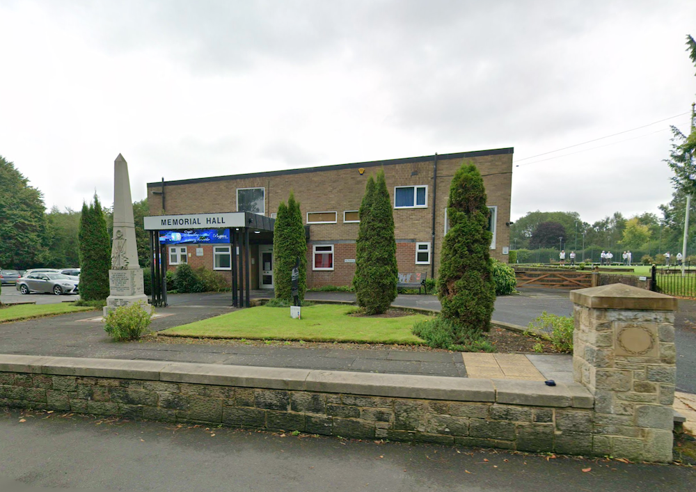 A brick memorial hall with a canopied entrance and a stone obelisk war memorial on the lawn in front