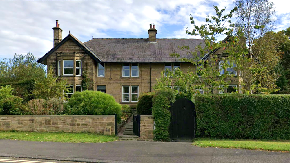 A pair of large stone Edwardian houses with a bay window and slate roof, behind a stone wall with iron gates