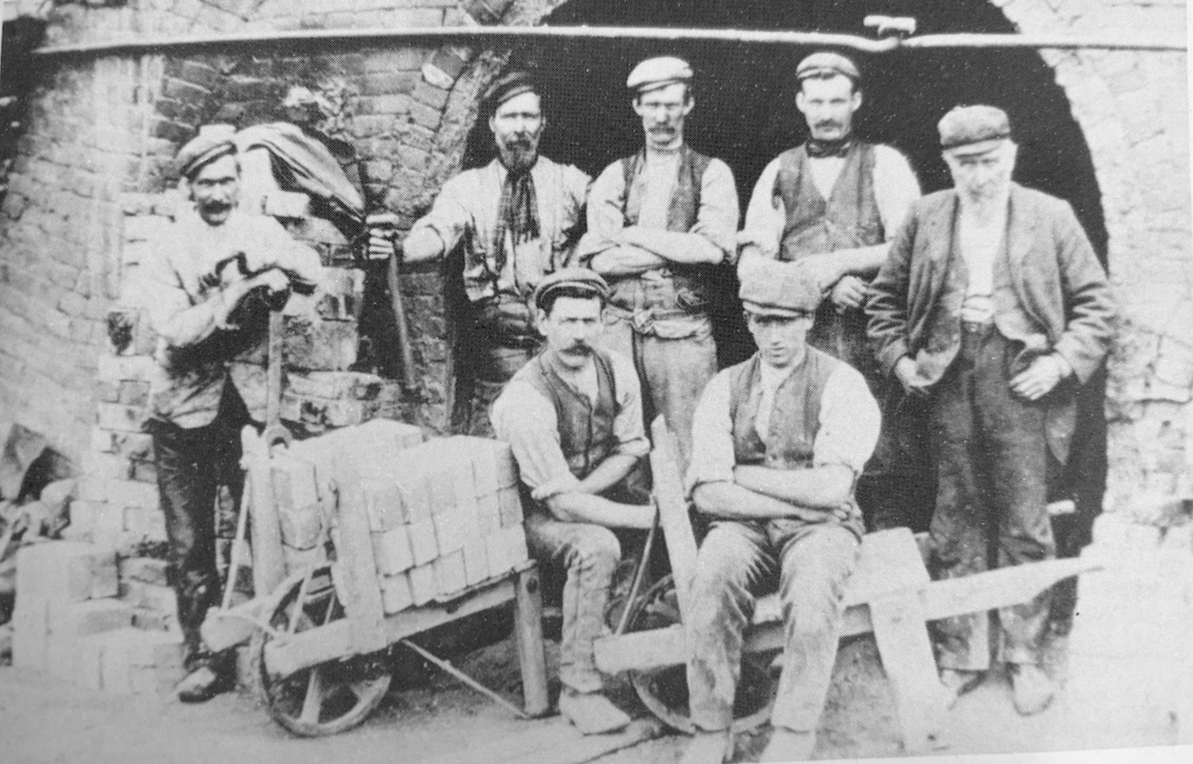 Black and white photograph of seven brickworks labourers posed in front of a kiln arch, with a barrow of freshly moulded bricks