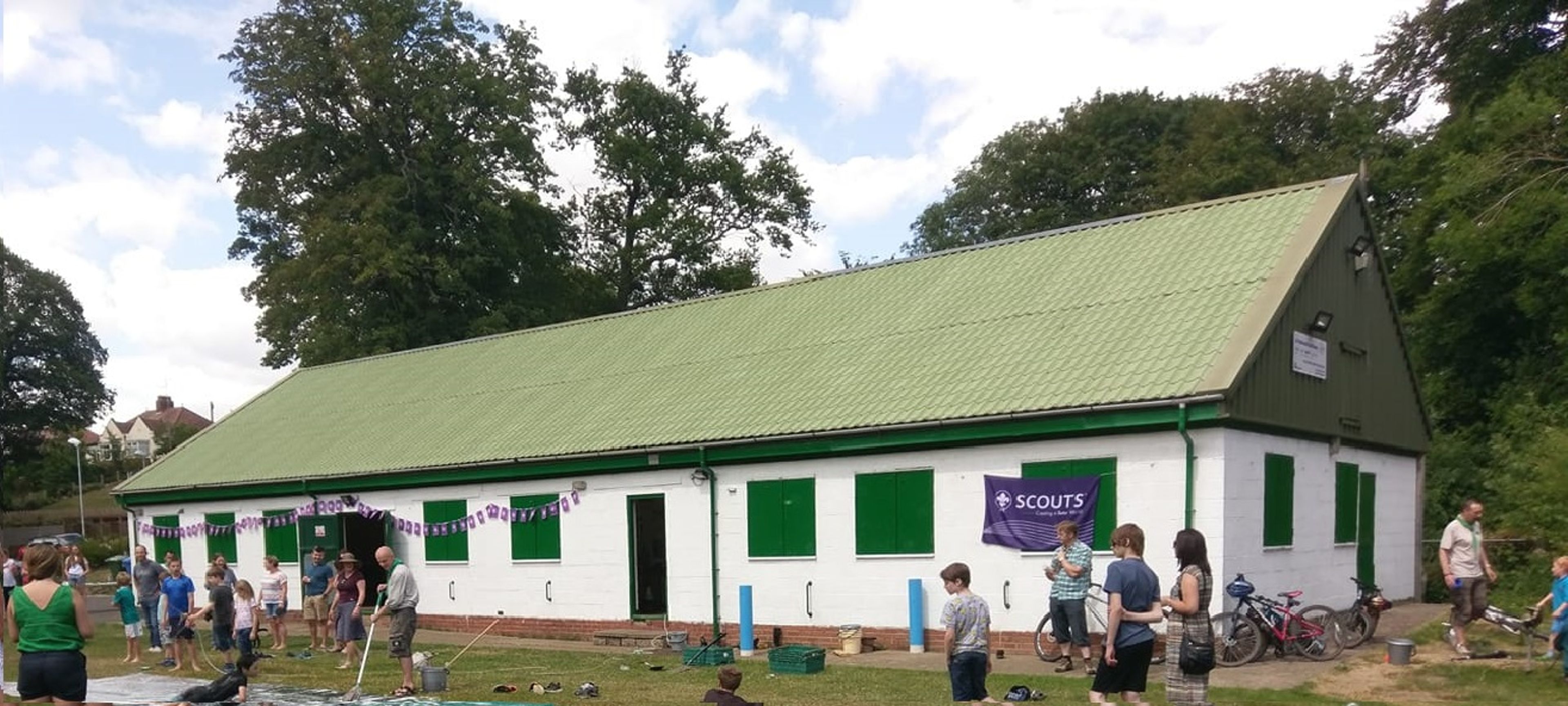 A long white scout hut with a green corrugated roof, bunting along the wall and people gathered outside on the grass