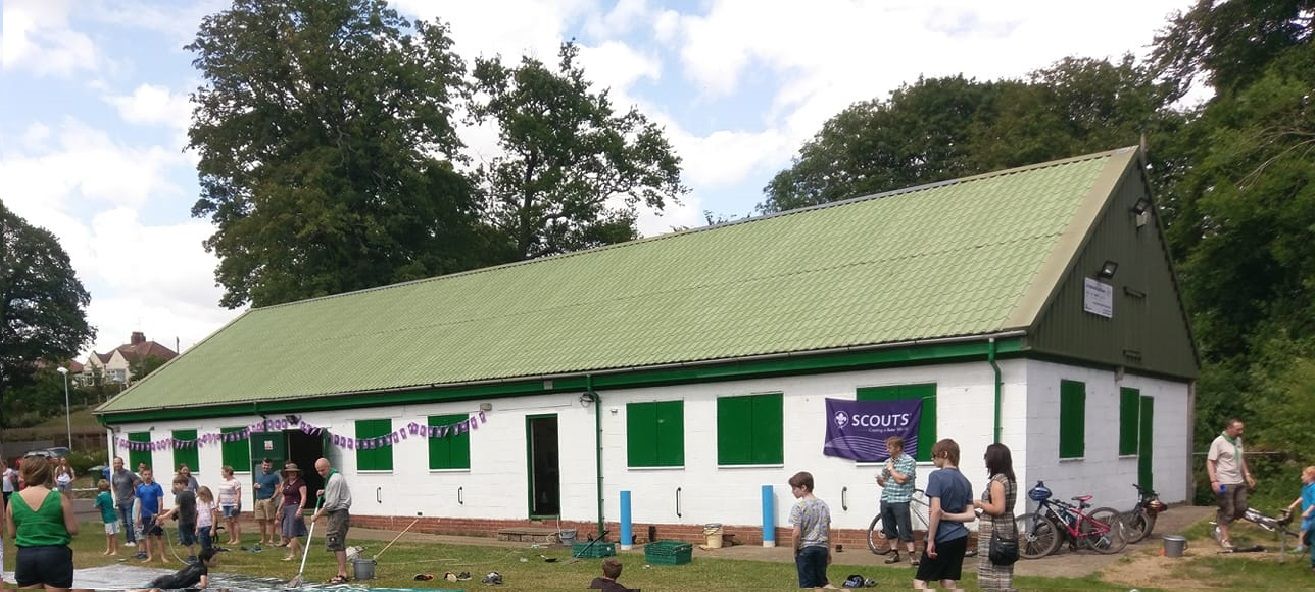 A long white scout hut with a green corrugated roof, bunting along the wall and people gathered outside on the grass