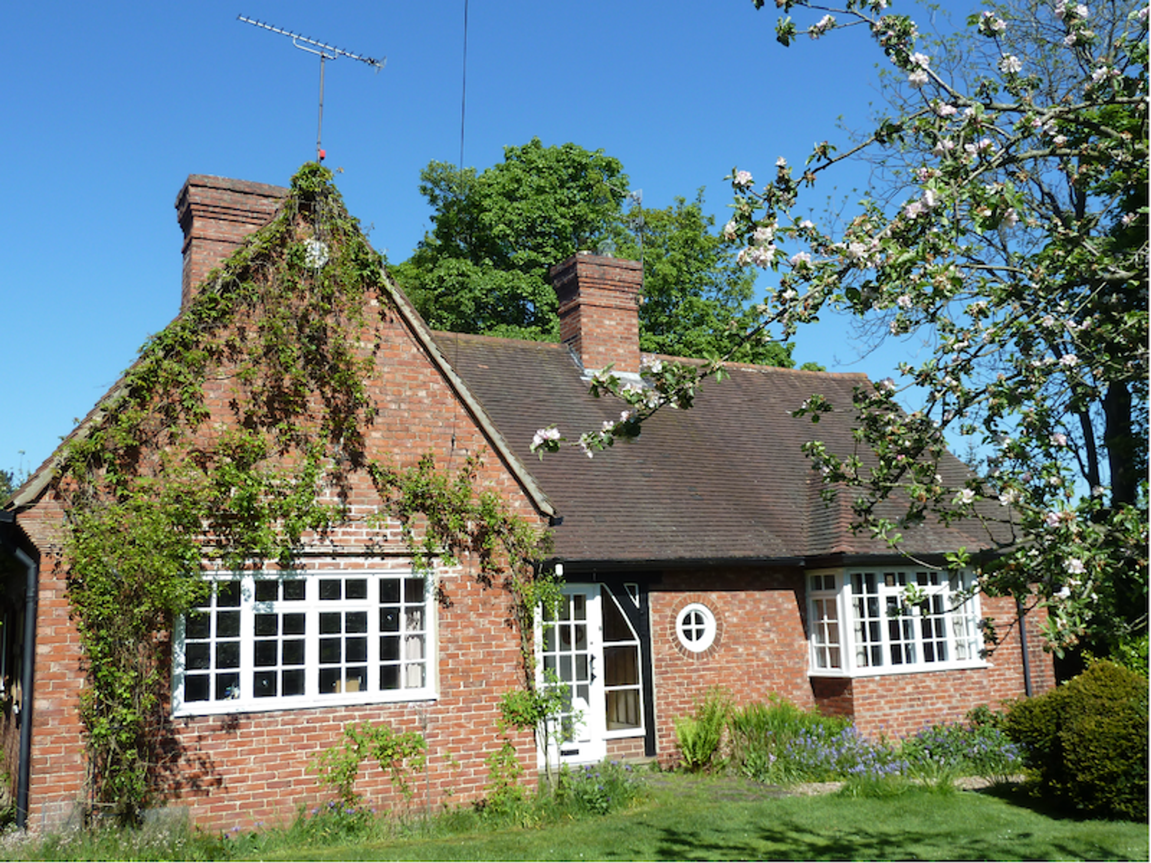 A red brick Arts and Crafts house with tall chimneys, a climbing rose across the gable, white casement windows and a circular window beside the door