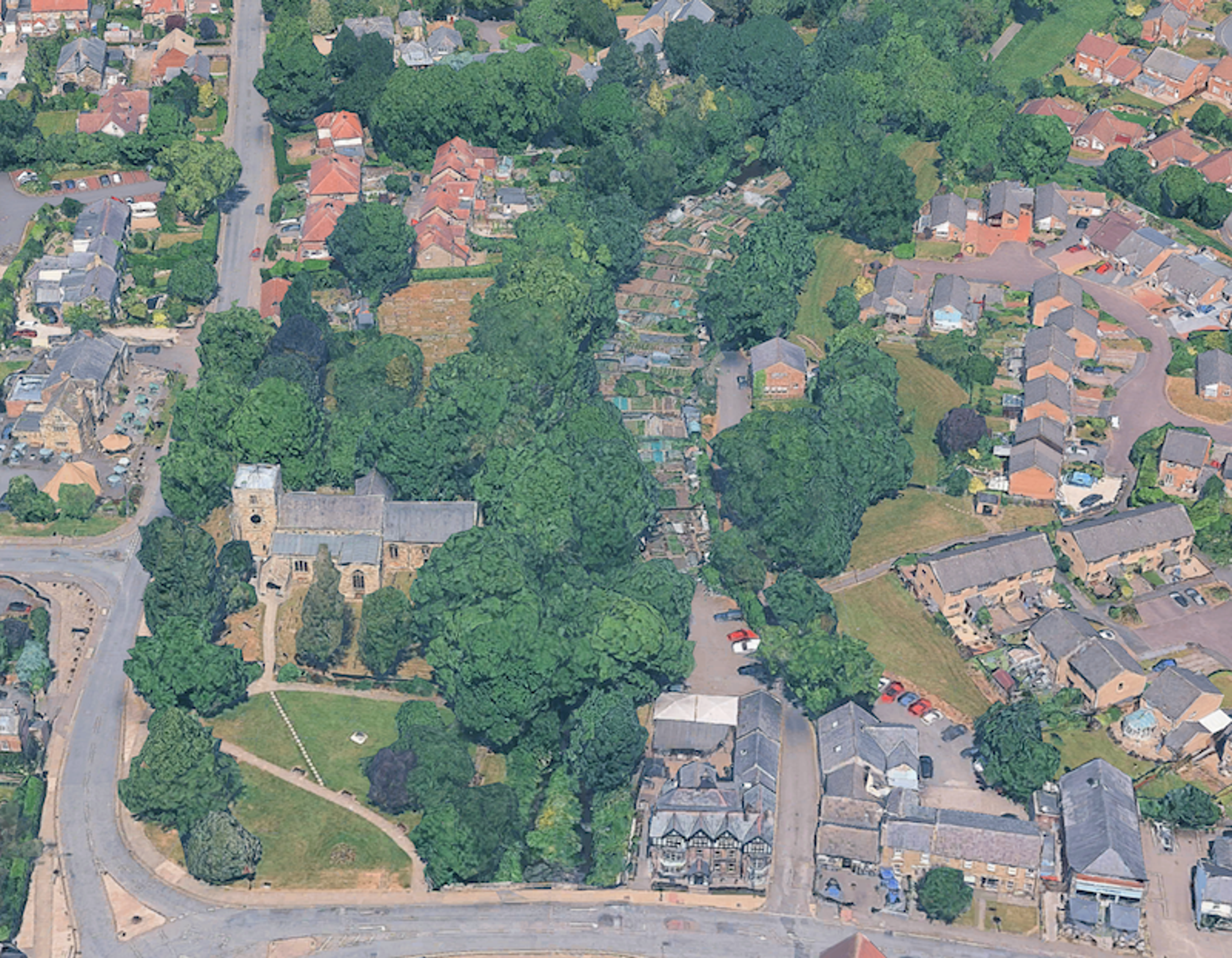 Aerial view of a village centre with a church among mature trees and a strip of allotments running behind the houses