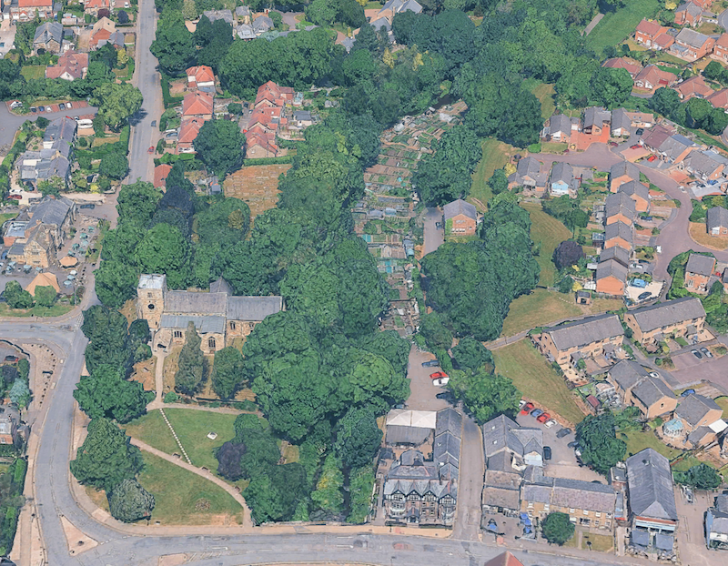Aerial view of a village centre with a church among mature trees and a strip of allotments running behind the houses