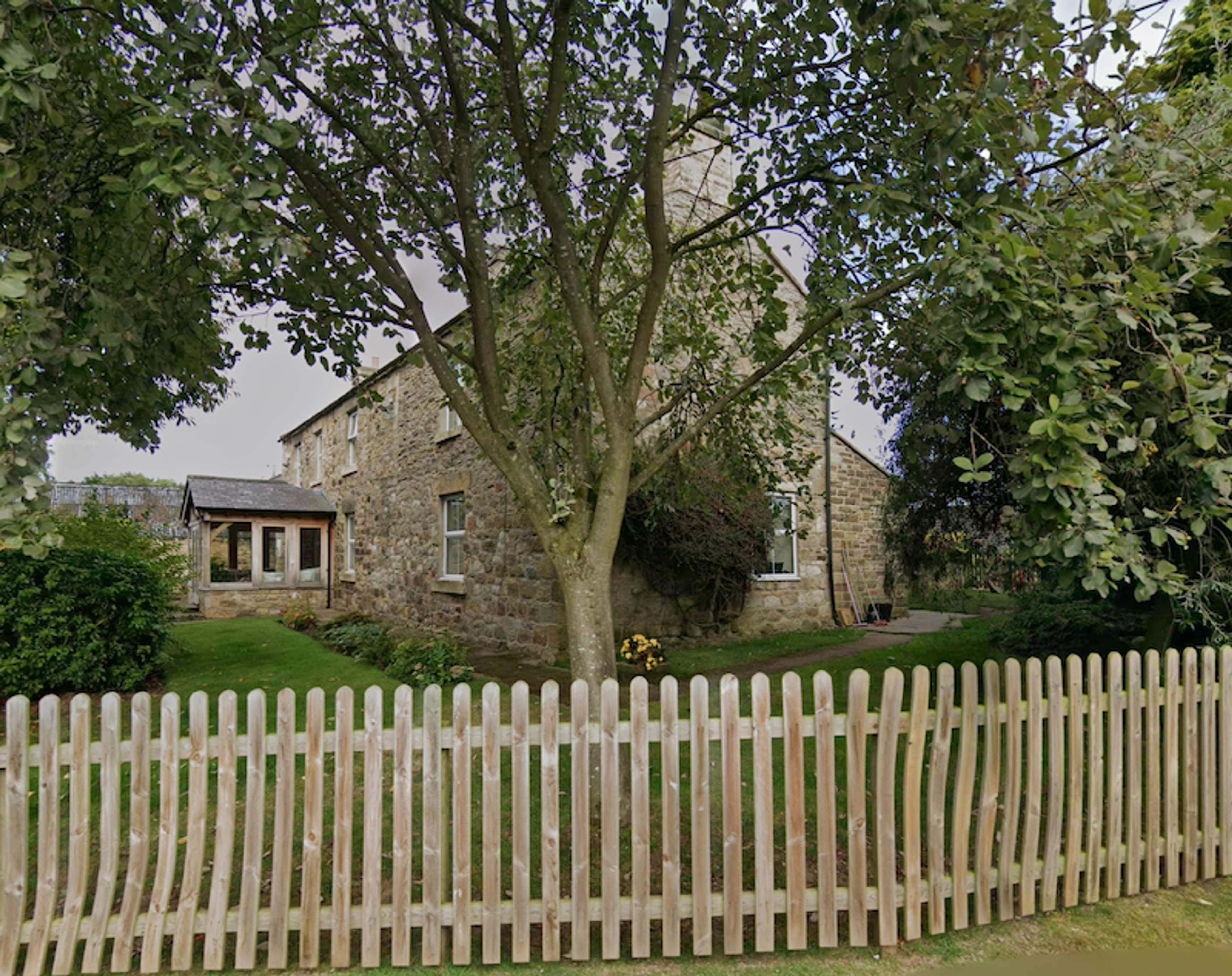 A stone farmhouse with a glazed porch behind a new wooden picket fence, a large tree in the front garden
