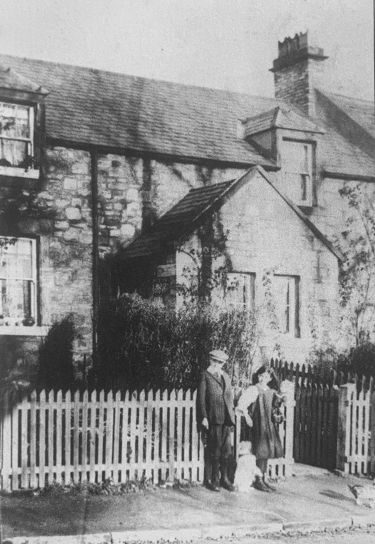Black and white photograph of a stone cottage terrace behind a white picket fence, with a man, woman and child standing at the gate