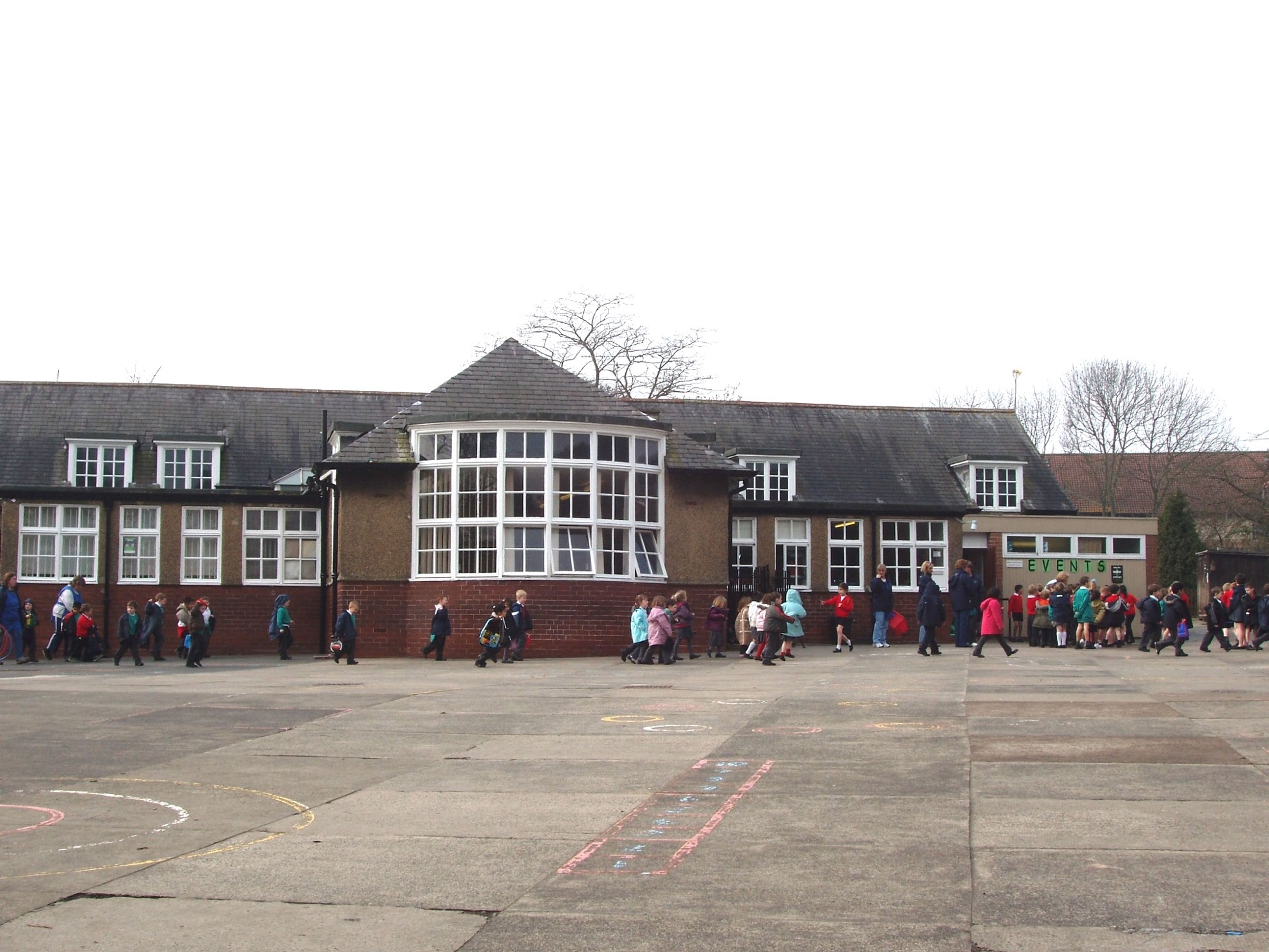 A single-storey school with a large bay window and hipped roof, children crossing the tarmac playground in front