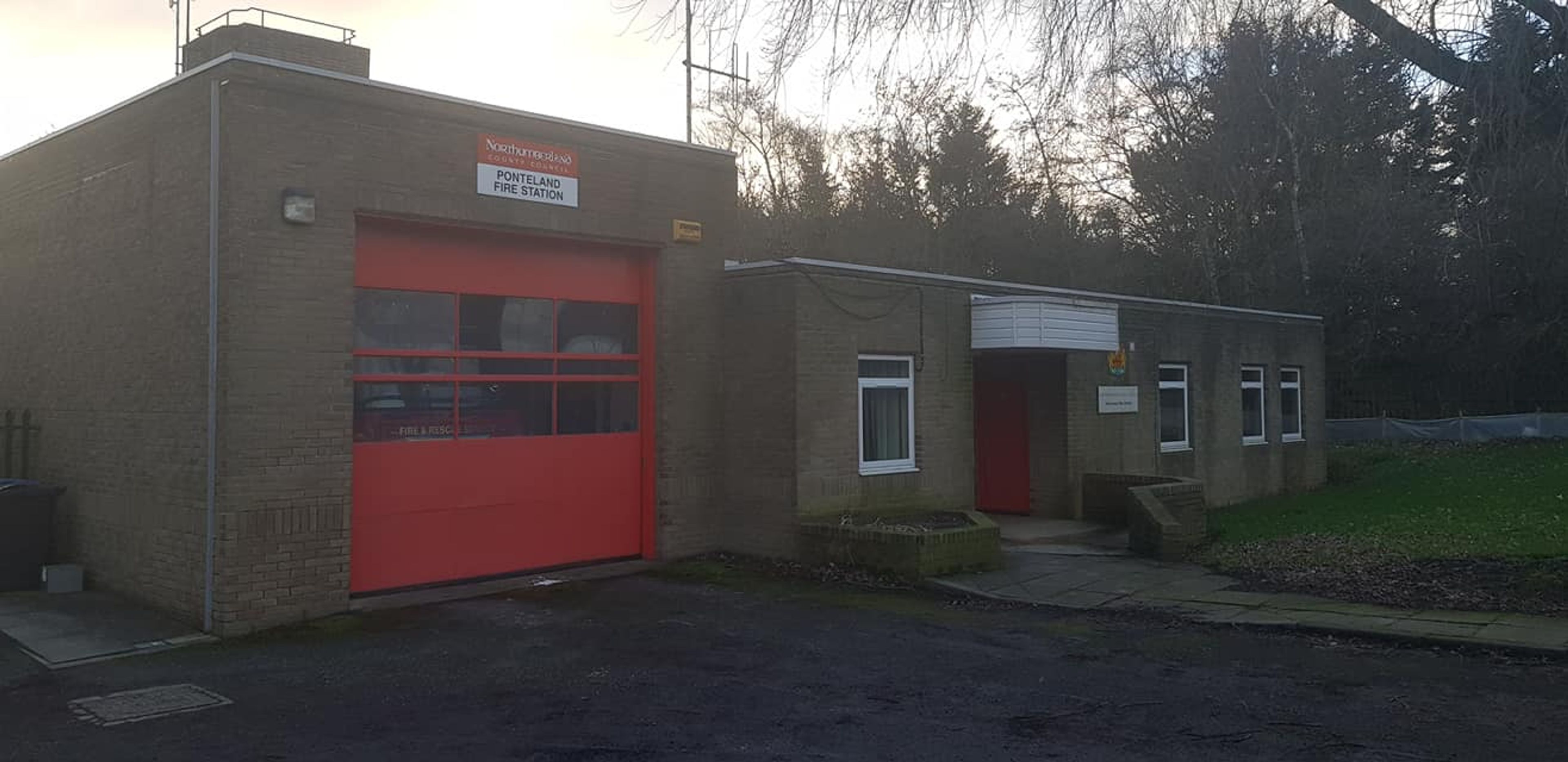 A small brick fire station with a red roller shutter door and an adjoining single-storey office block
