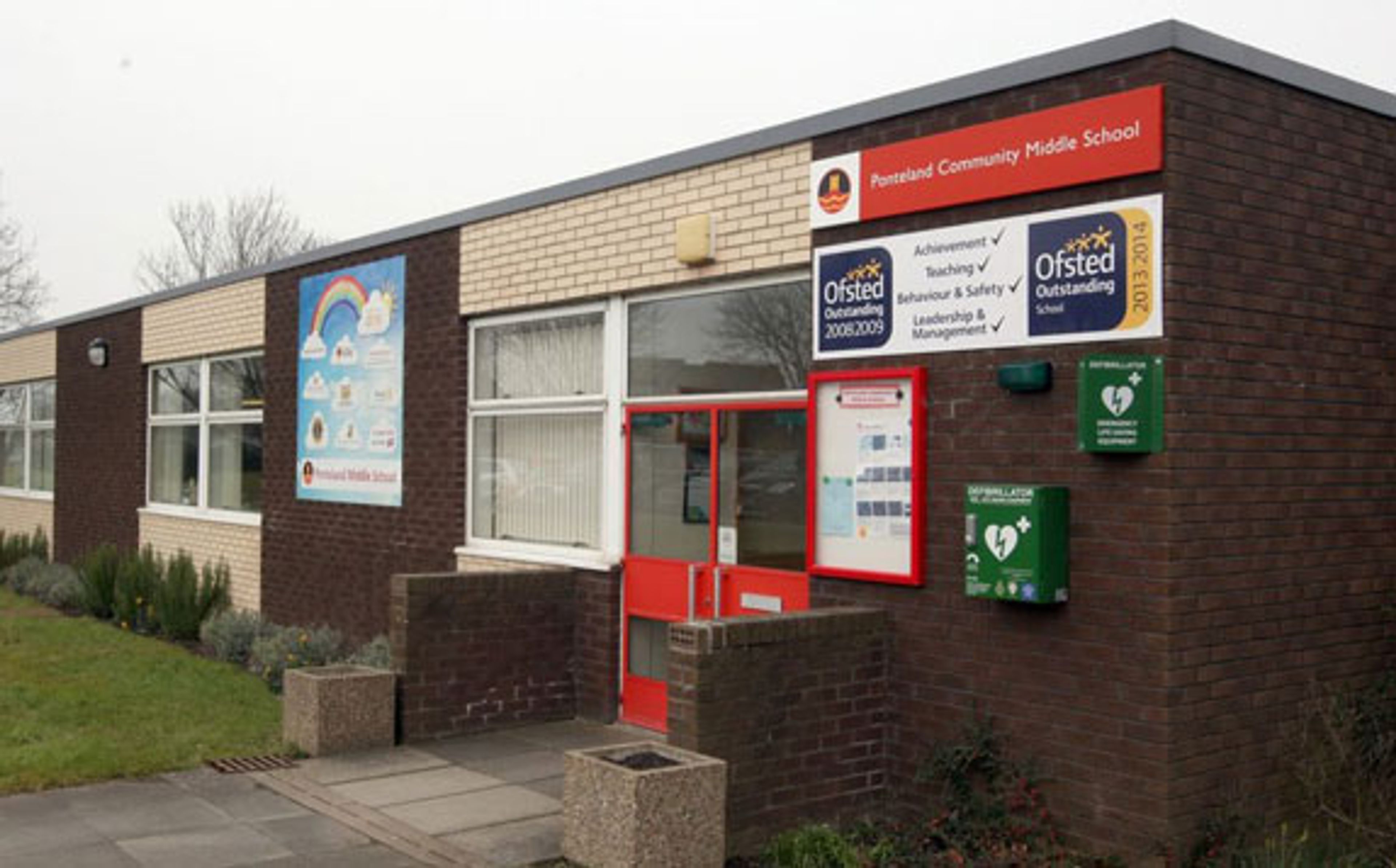 A single-storey brick school entrance with red doors, noticeboards and Ofsted Outstanding banners on the wall